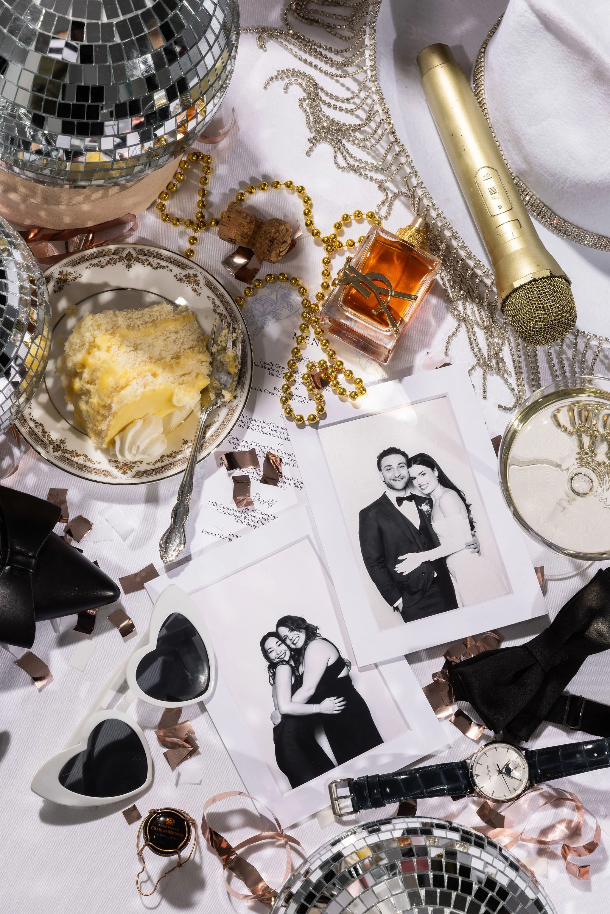 A collection of wedding or celebration items on a white tablecloth, including black and white photos of a couple, a slice of cake, desserts, a microphone, sunglasses, a watch, and decorative disco balls, with gold and rose gold bead garlands and ribb