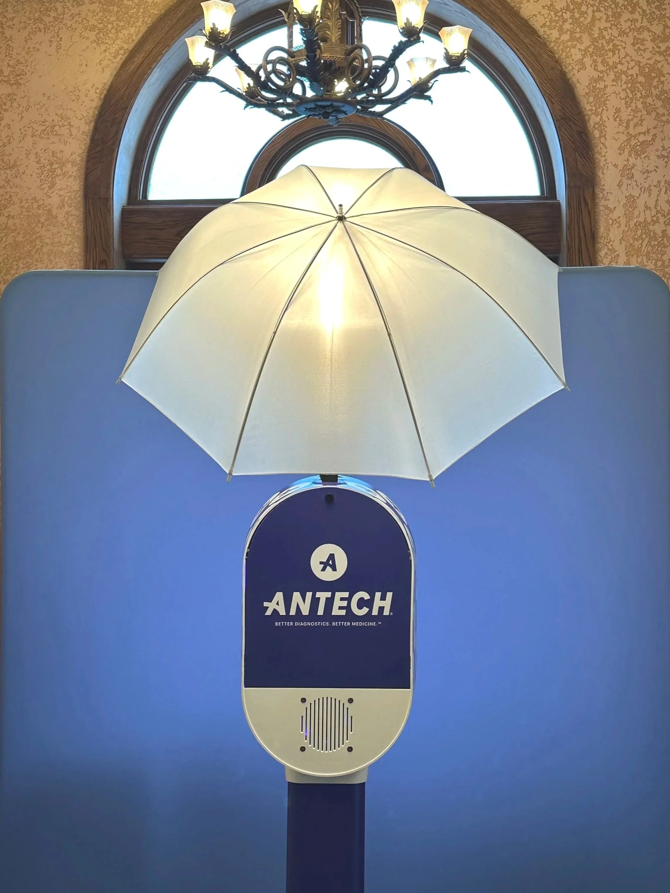 A photo booth vinyl wrapped with Antech branding set up at a conference, partition, with a chandelier and arched window in the background.