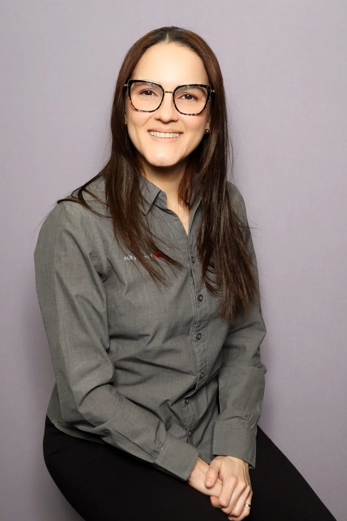 A woman with glasses and long brown hair, wearing a gray button-up shirt, smiling and seated against a plain gray background.