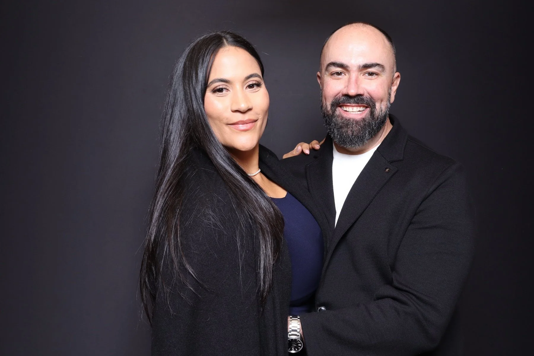A smiling man with a beard and a woman with long dark hair posing together against a dark background.