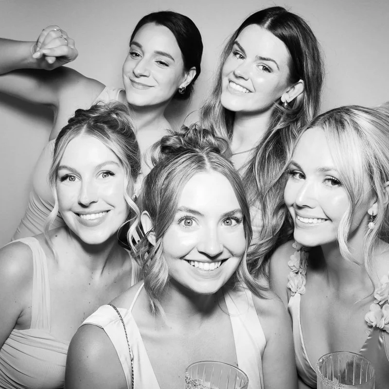 Group of six women smiling and posing for a black-and-white photo, some with drinks in hand, wearing light-colored dresses.