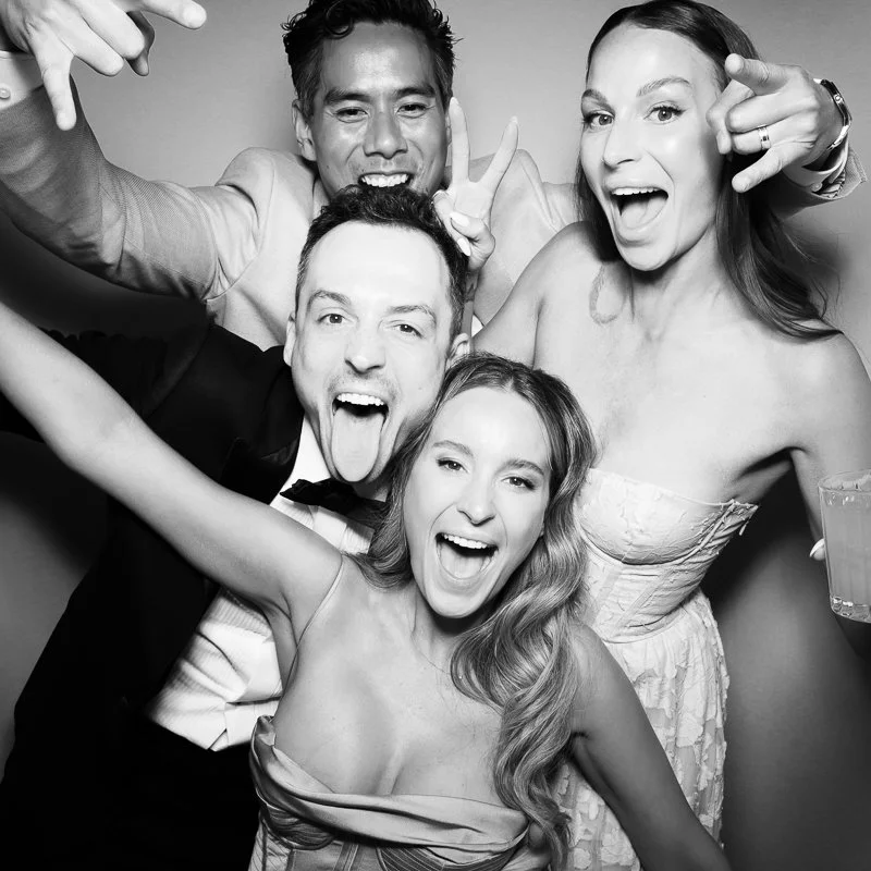Four people celebrating at a party, taking a selfie. They are smiling and making playful gestures.