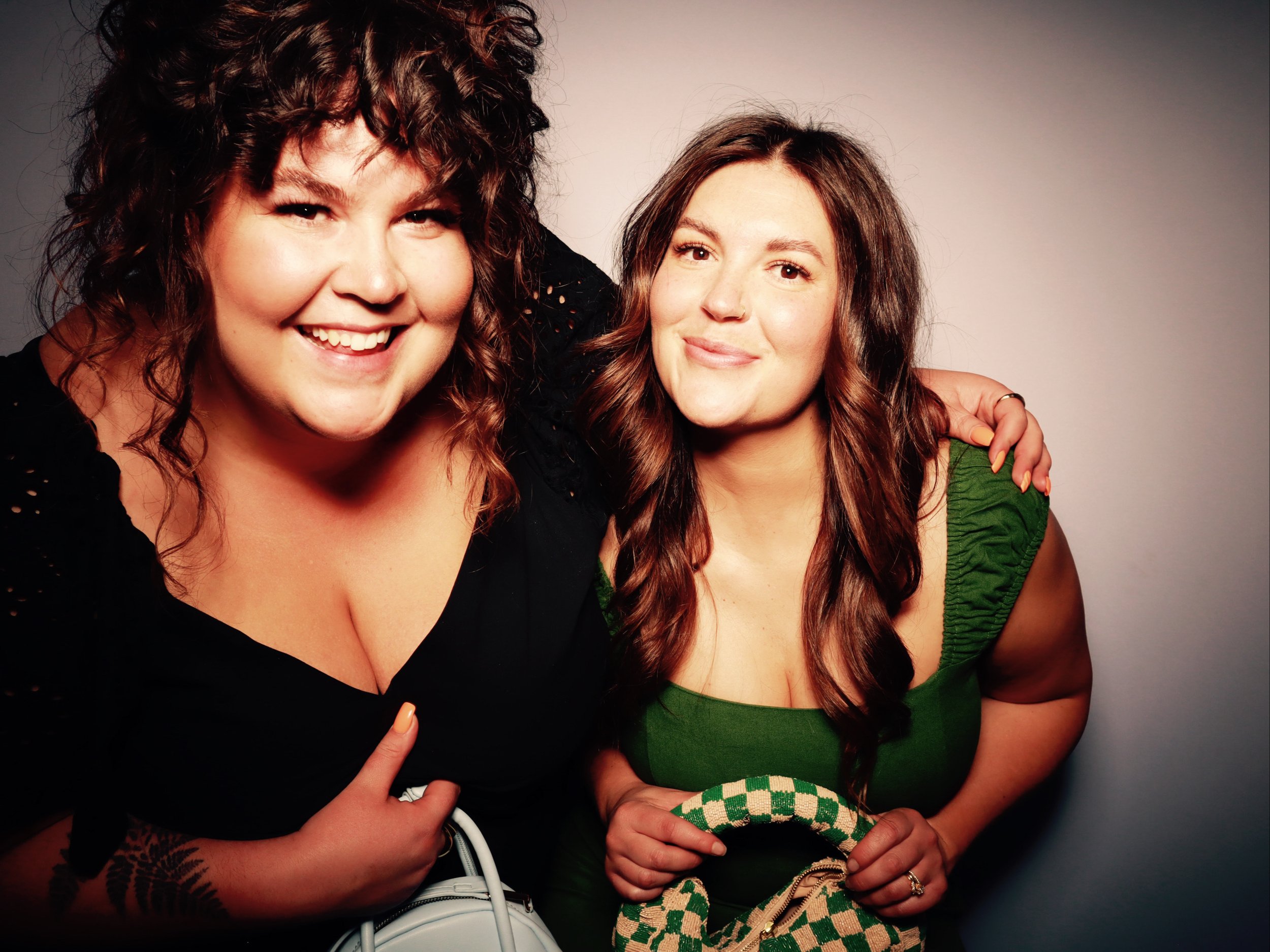 Two women smiling, one with curly dark hair in a black top, and the other with straight brown hair in a green top, standing closely with arms around each other against a plain gray background.
