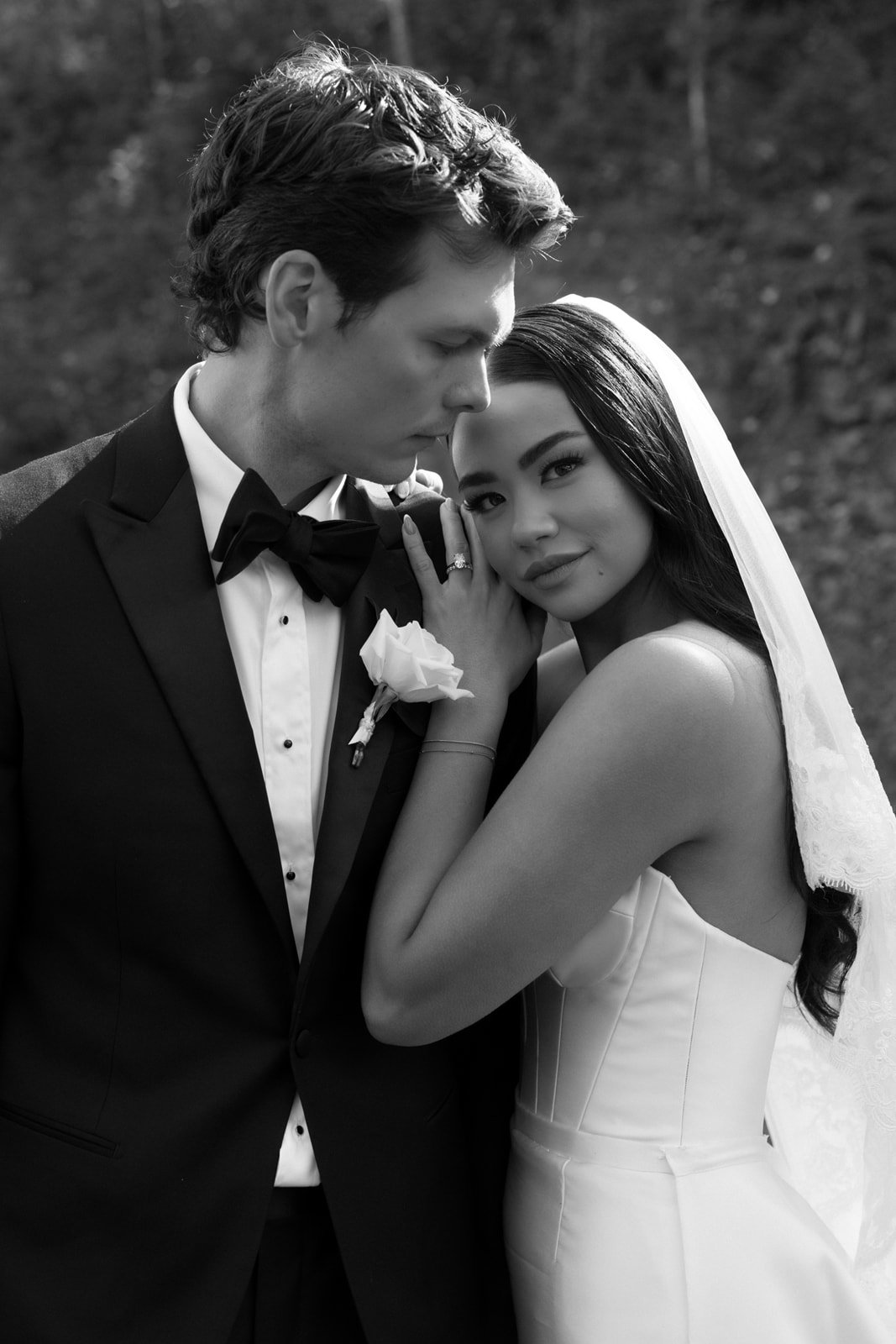 Black and white photo of a bride and groom on their wedding day, standing close together outdoors. The groom is in a tuxedo with a bow tie and a boutonnière, with his eyes closed leaning his forehead against the bride's. The bride, with long dark hai