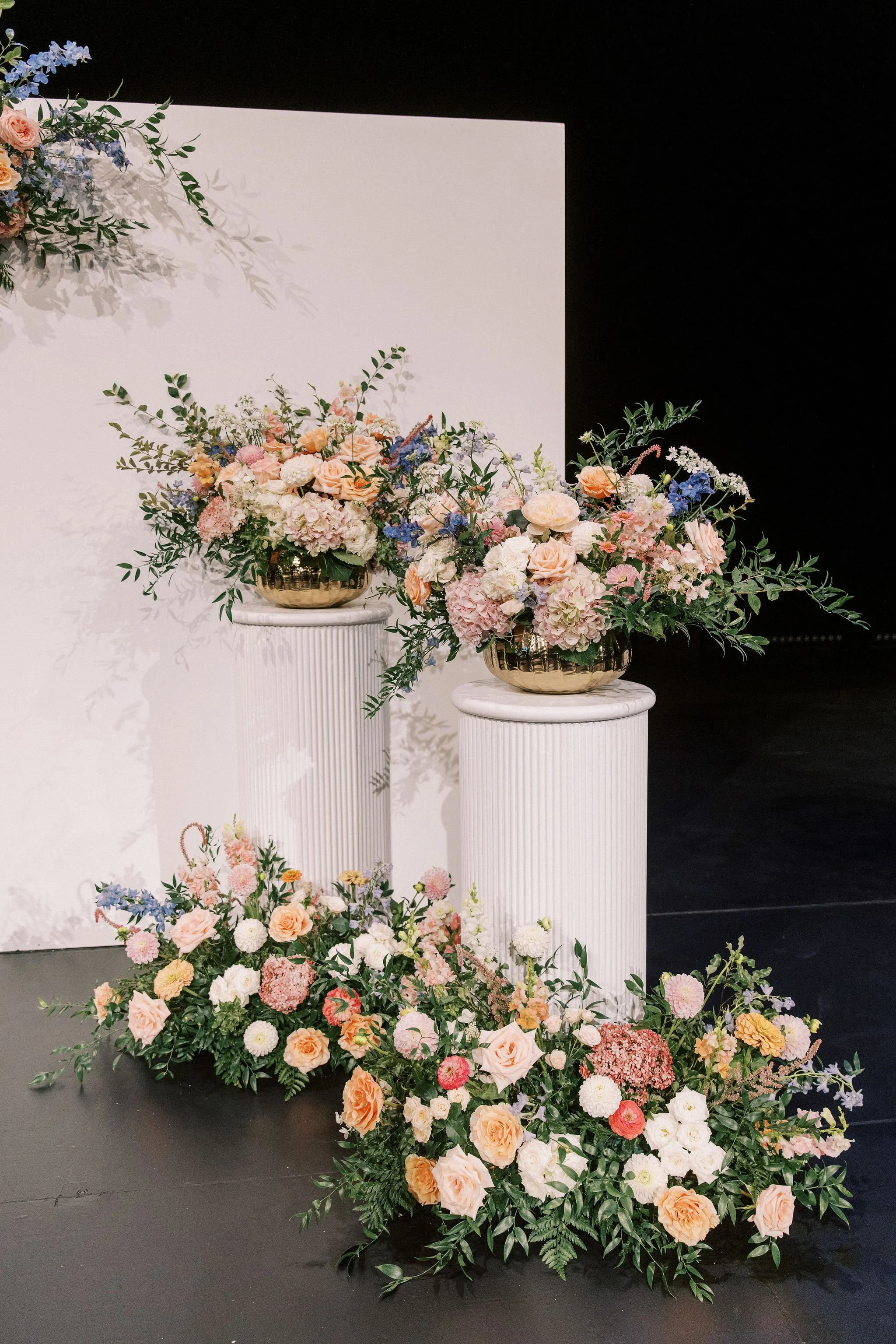 Arrangement of pastel-colored flowers, including roses and hydrangeas, in gold bowls on white pedestals and a floral display on the floor against a black background.