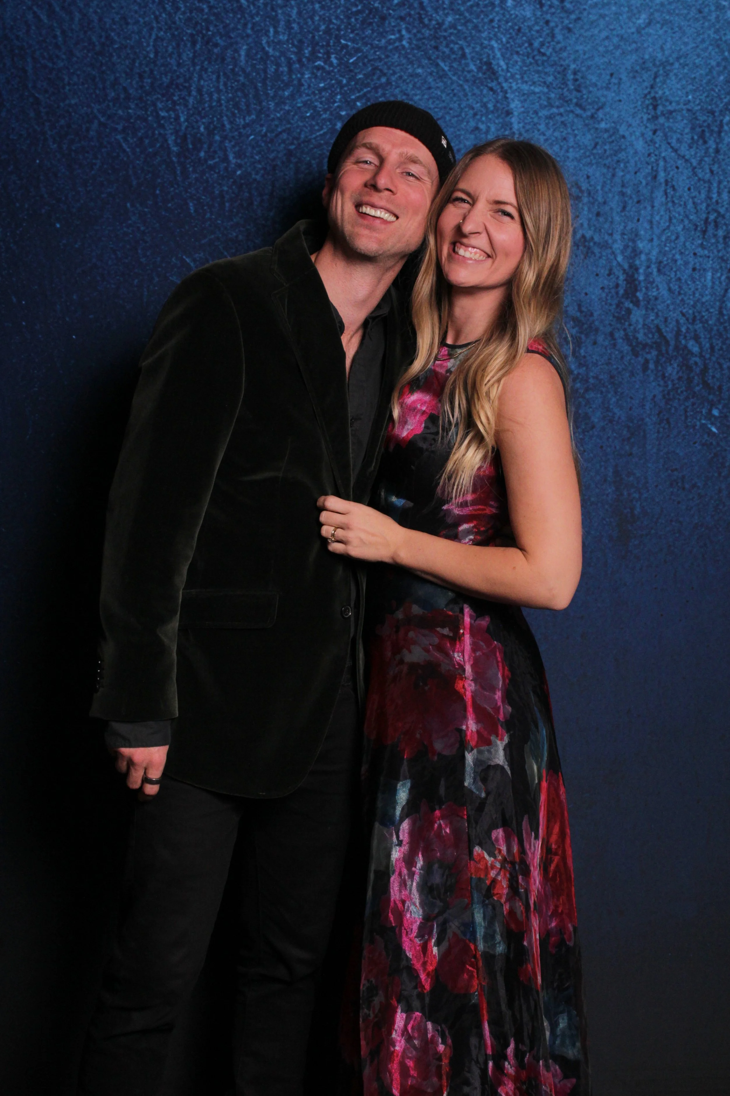 A man and woman posing together against a blue textured background, smiling and showing affection.
