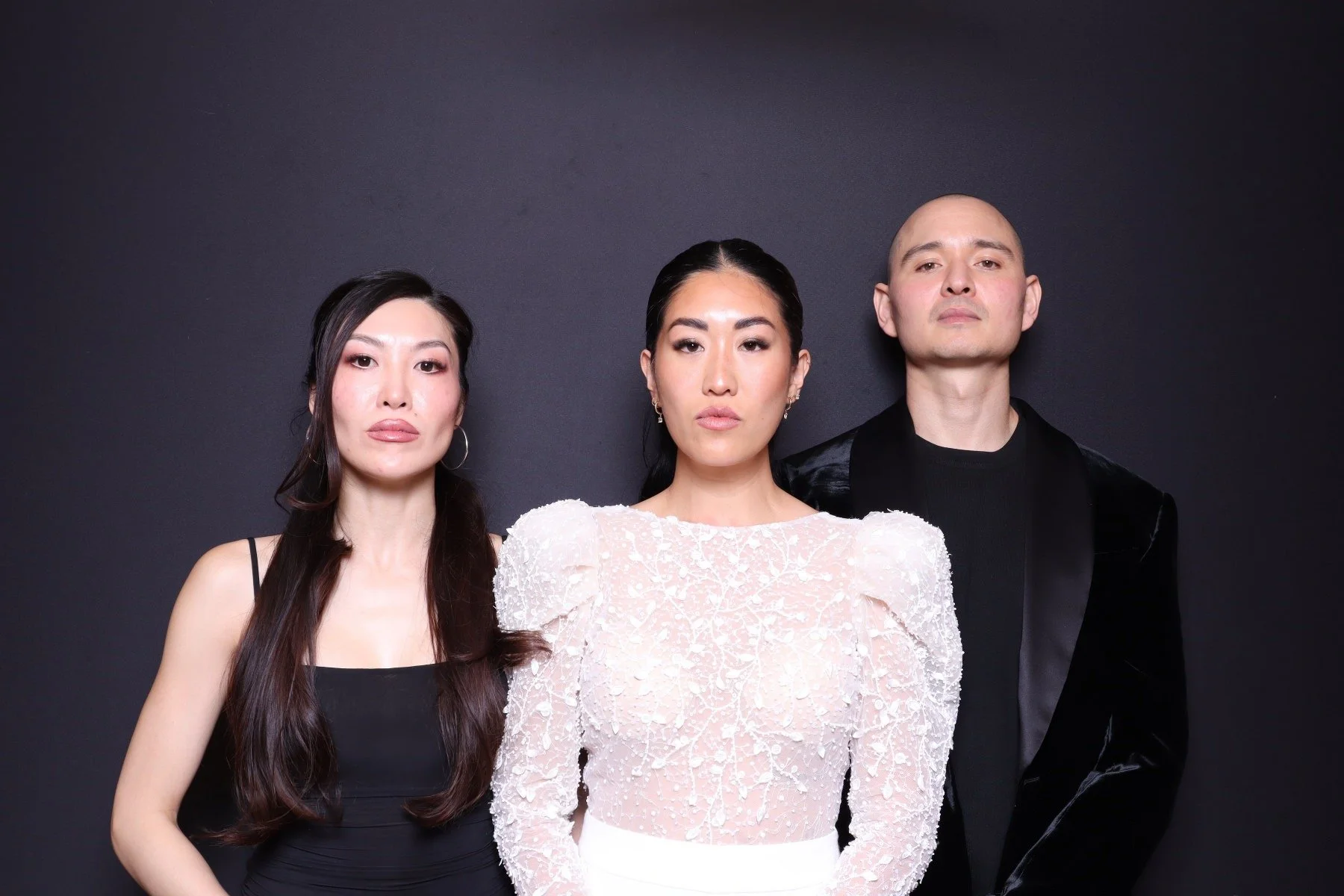 Three people standing against a black background. Two women and one man, dressed in stylish formal attire, looking directly at the camera with serious expressions.