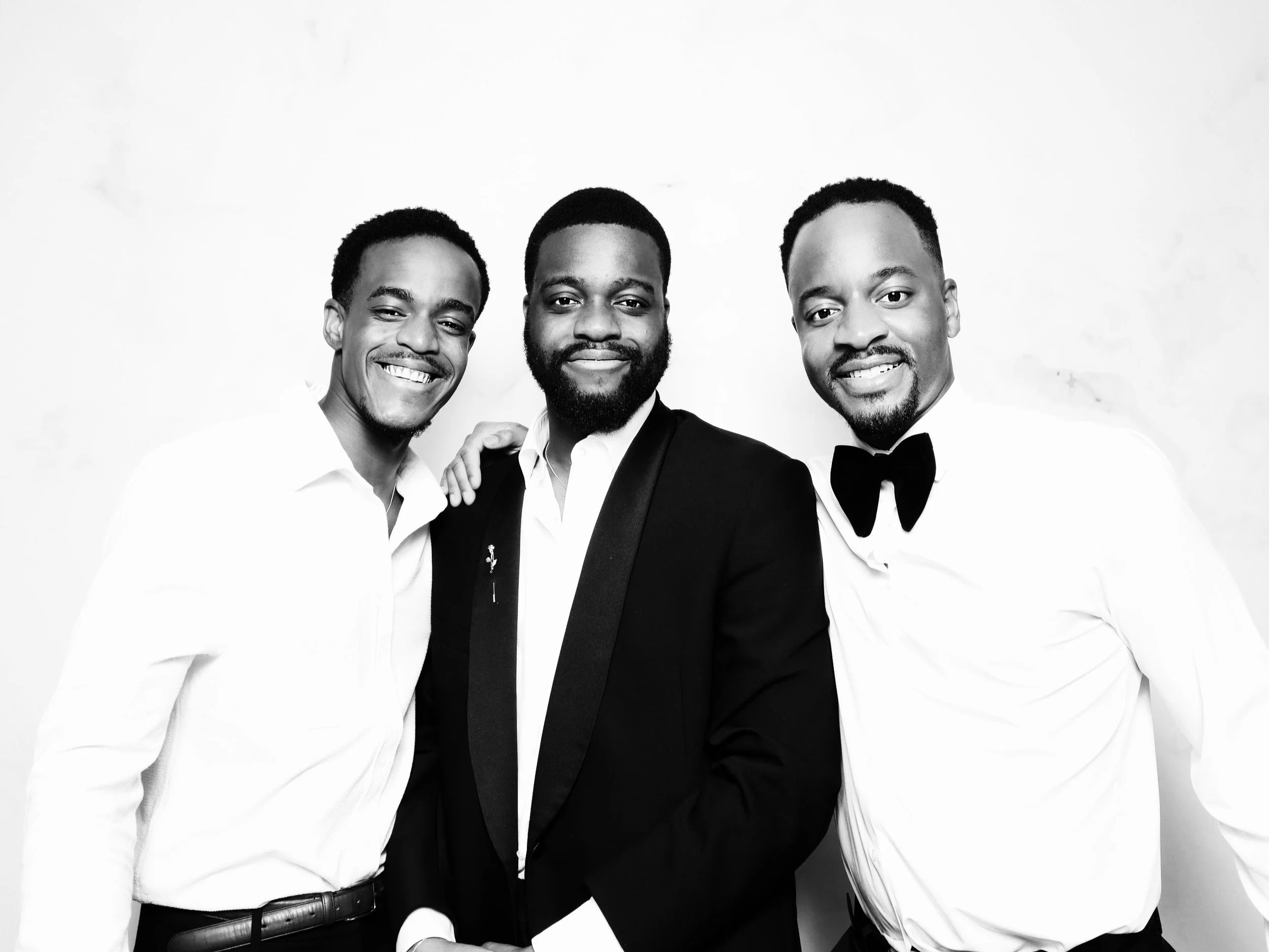 Three men dressed in formal attire, smiling and standing close together against a plain background.