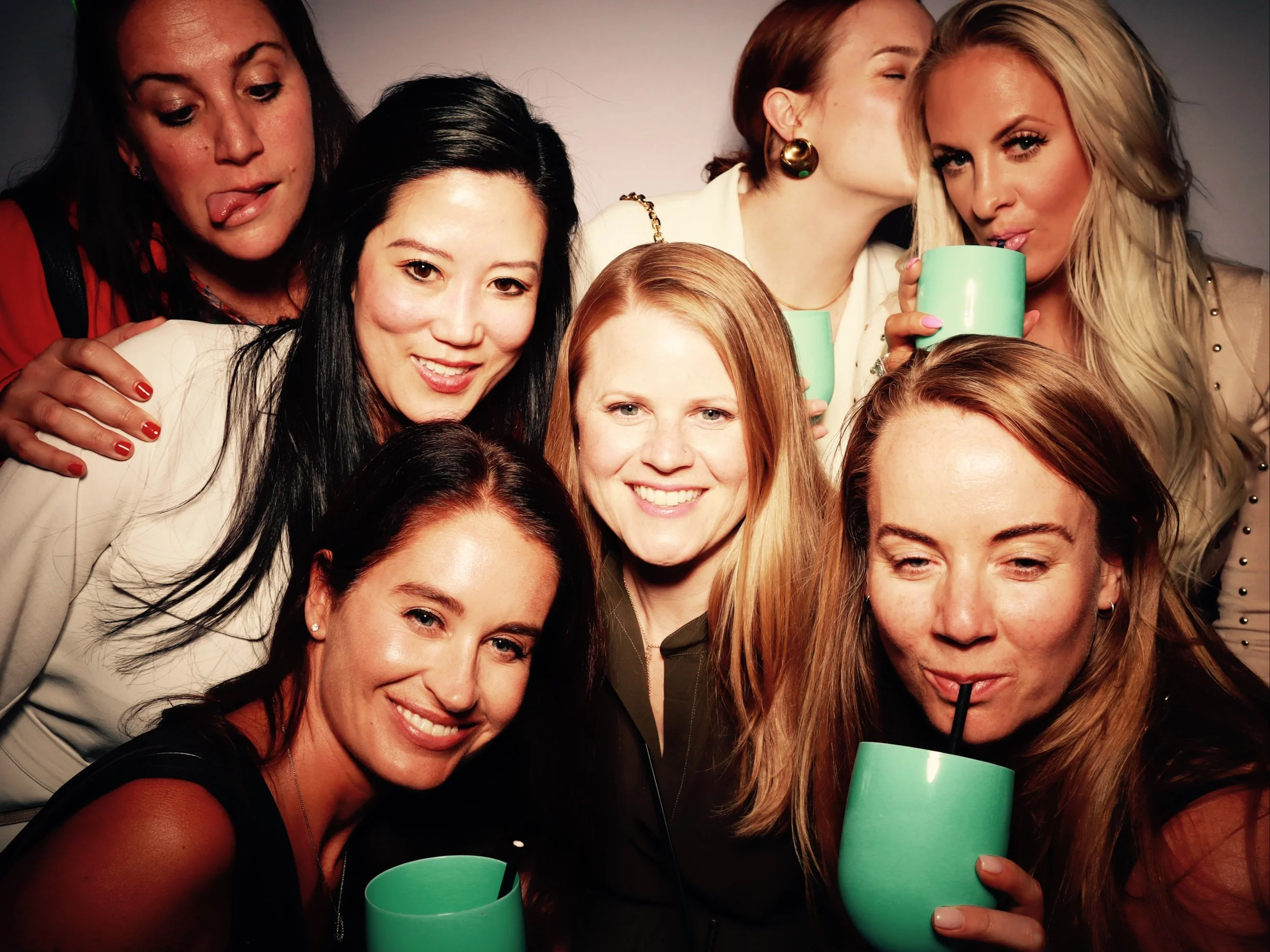 Eight women in a group photo, some holding turquoise mugs, some drinking through straws, smiling and enjoying a social gathering or party.