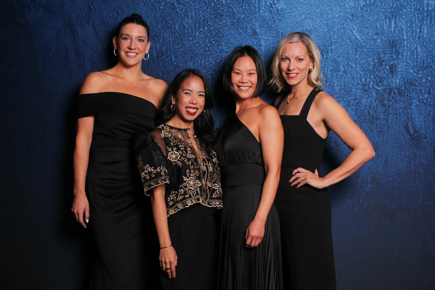 Four women in black dresses standing against a blue textured wall, smiling at the camera.