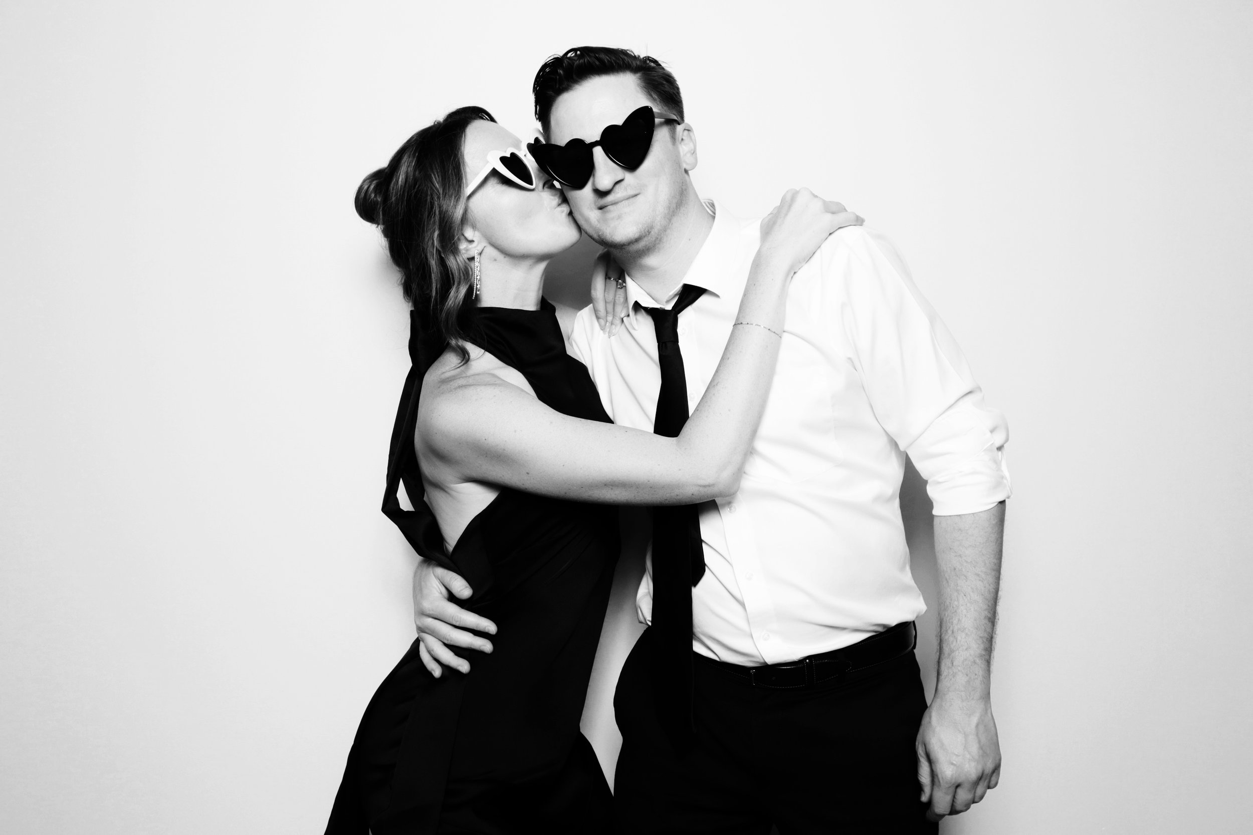 A woman in a black dress kissing a man on the cheek, both wearing heart-shaped sunglasses, posing against a plain background.