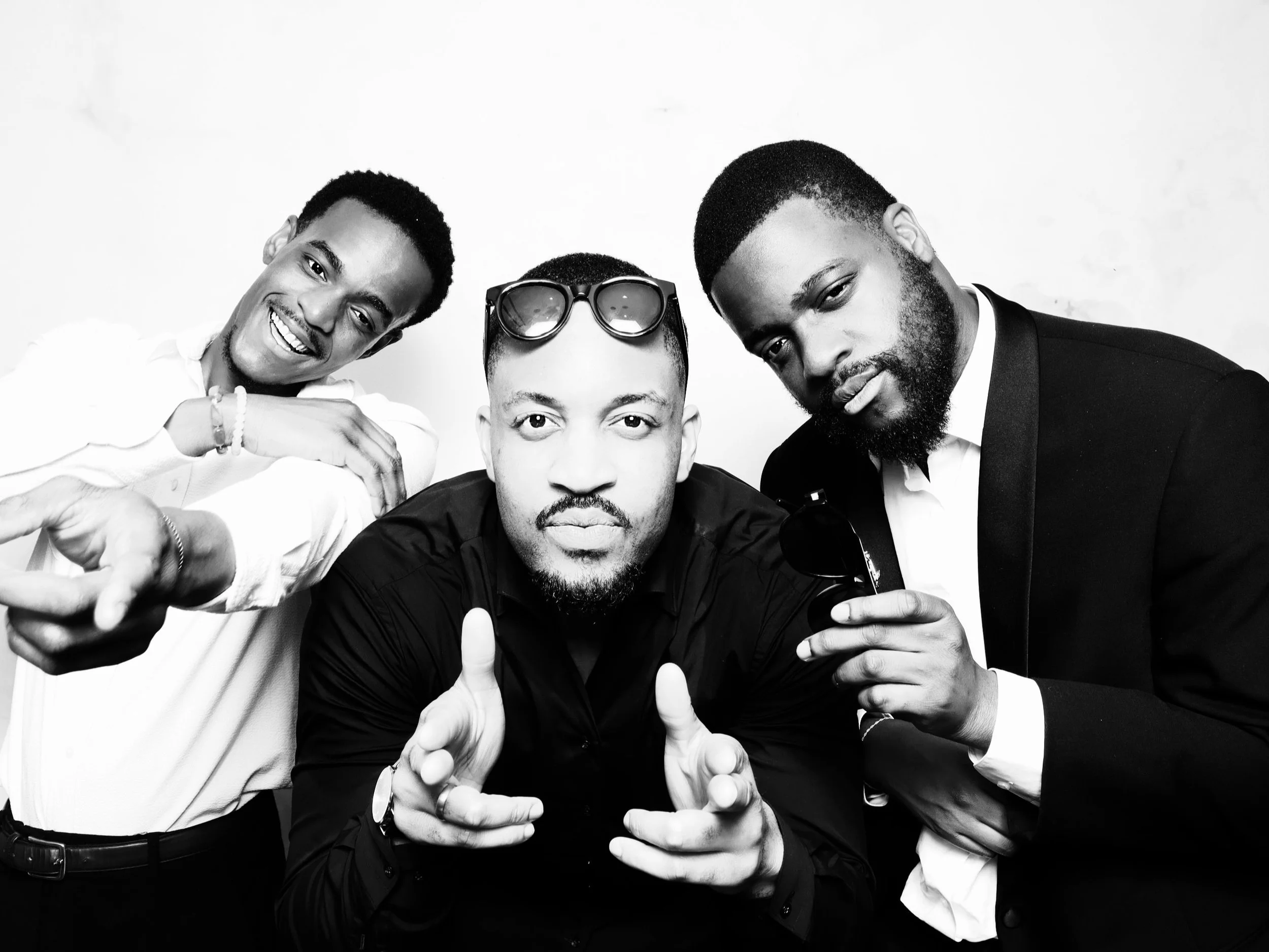 Black and white photo of three men posing together, two wearing suits and one wearing a casual shirt, with one holding sunglasses.