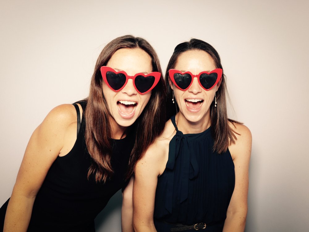 Two women wearing black tops and red heart-shaped sunglasses, smiling and laughing in front of a plain light-colored background.