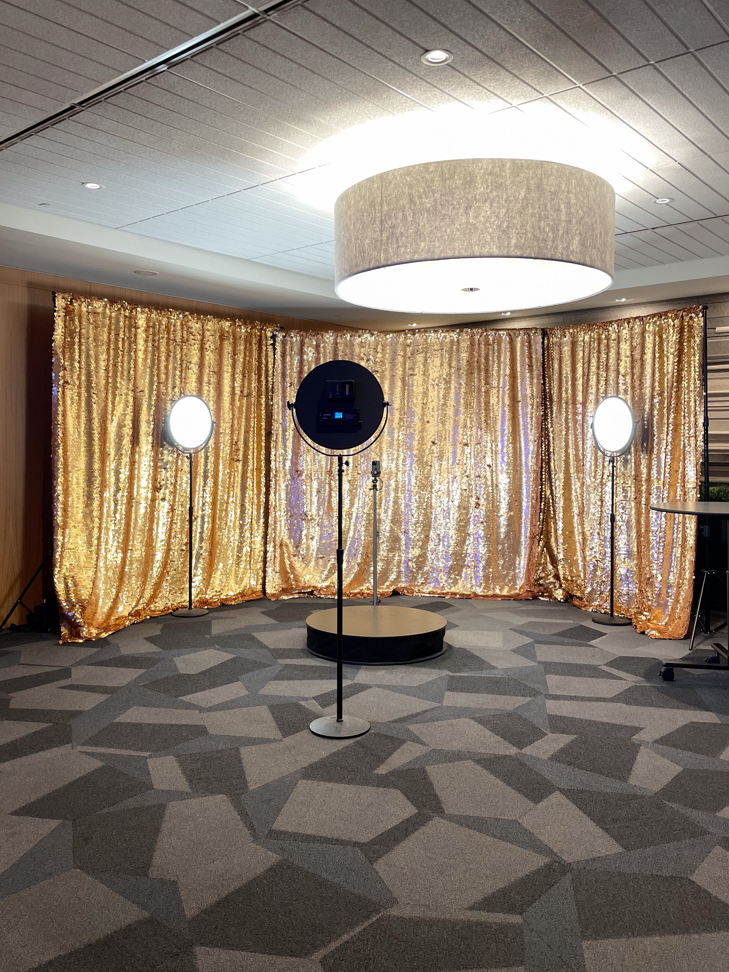 Photo of a photo booth setup with three circular lights and a golden sequin backdrop, set on a patterned carpeted floor.