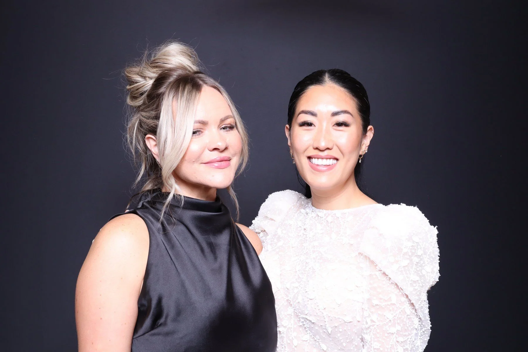 Two women smiling, one with blonde hair in a black sleeveless dress, and the other with black hair in a white textured dress, standing against a dark background.