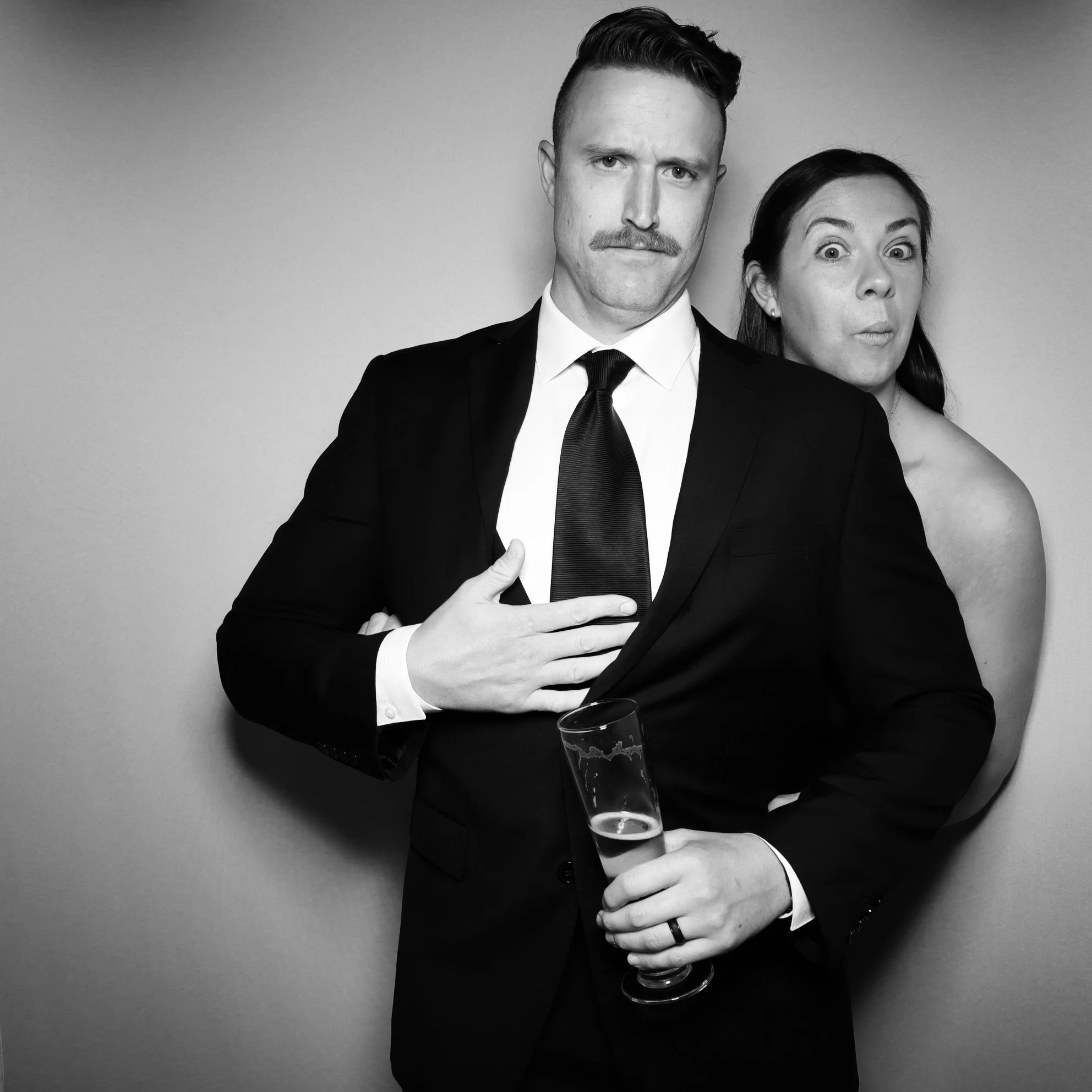Black and white photo of a man in a suit holding a champagne glass, with a woman standing behind him making a surprised face.