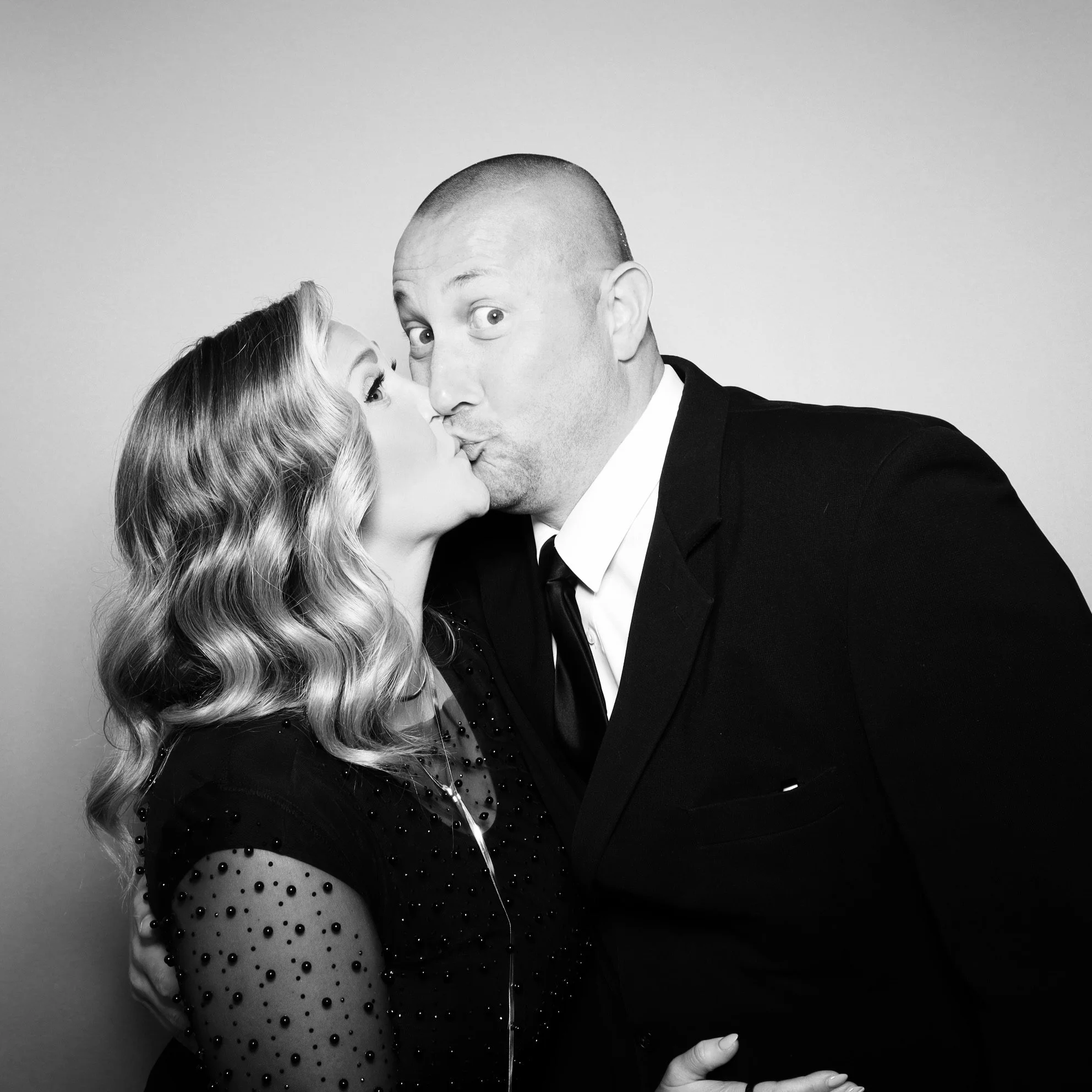 A woman with wavy blonde hair and a man with a shaved head kissing on the cheek, both dressed formally against a plain background.