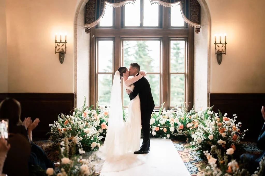 A bride and groom kissing during a wedding ceremony, standing on a white aisle surrounded by floral arrangements, in front of large window with curtains in an elegant venue.