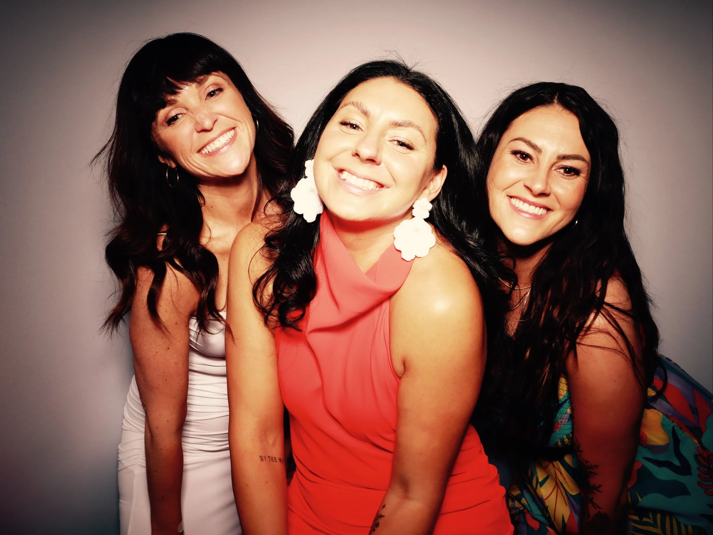 Three women smiling and posing together against a plain background. The woman in the center is wearing a coral dress with large white earrings, and the women on either side are wearing dresses with darker hairstyles.