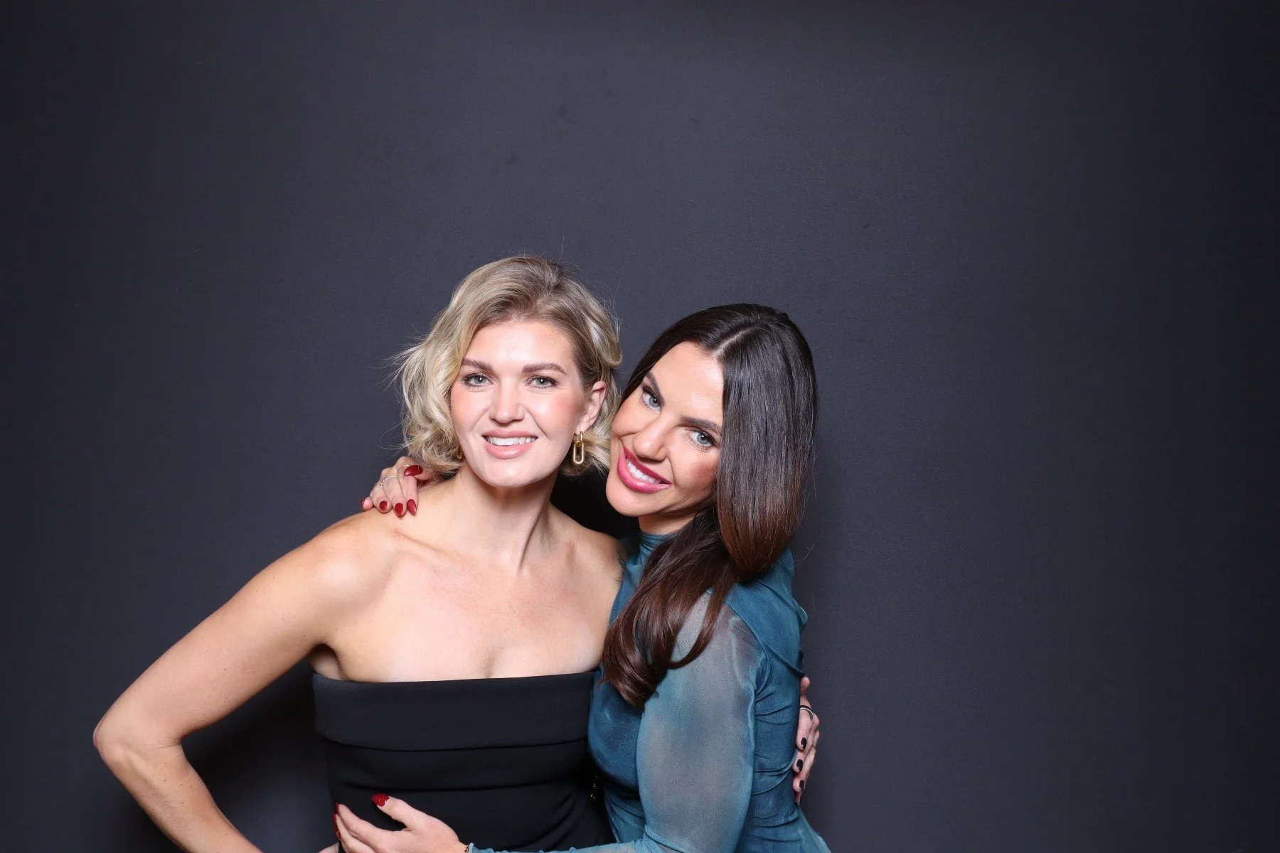 Two women hugging and smiling against a black background, one with blonde hair in a black strapless dress, the other with long dark hair in a blue top.