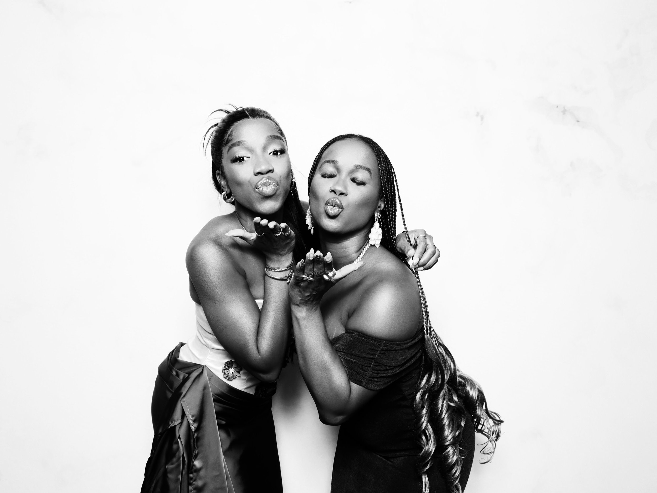 Two women in elegant dresses blowing kisses toward the camera, standing close together against a plain white background.