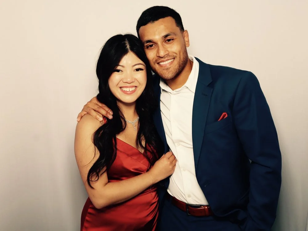 A smiling woman in a red dress with black hair and a man in a blue suit and white shirt smiling together against a plain light-colored background.