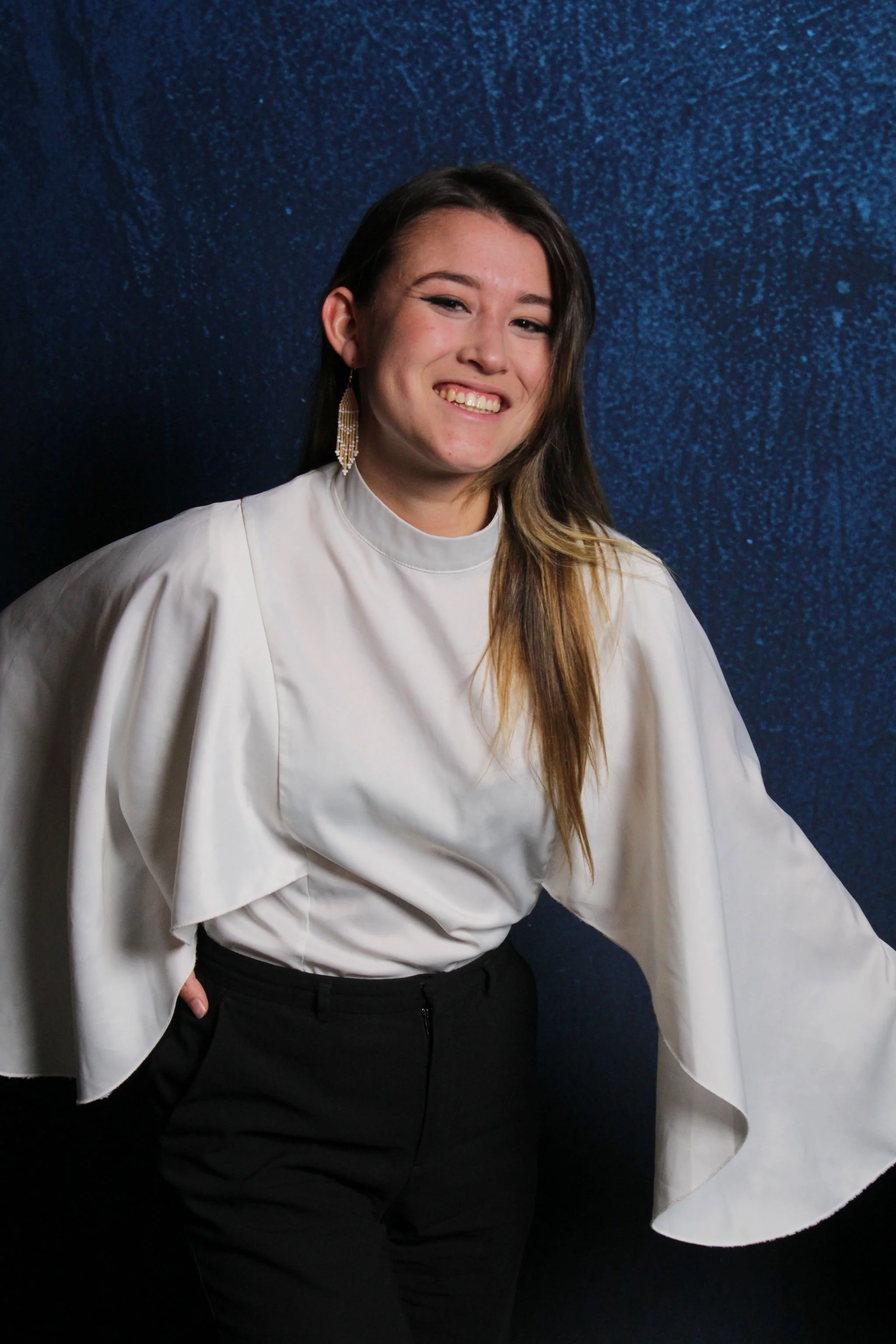 A woman with long brown hair, smiling, wearing a white blouse with dramatic sleeves, black pants, and gold earrings, posing against a blue background.