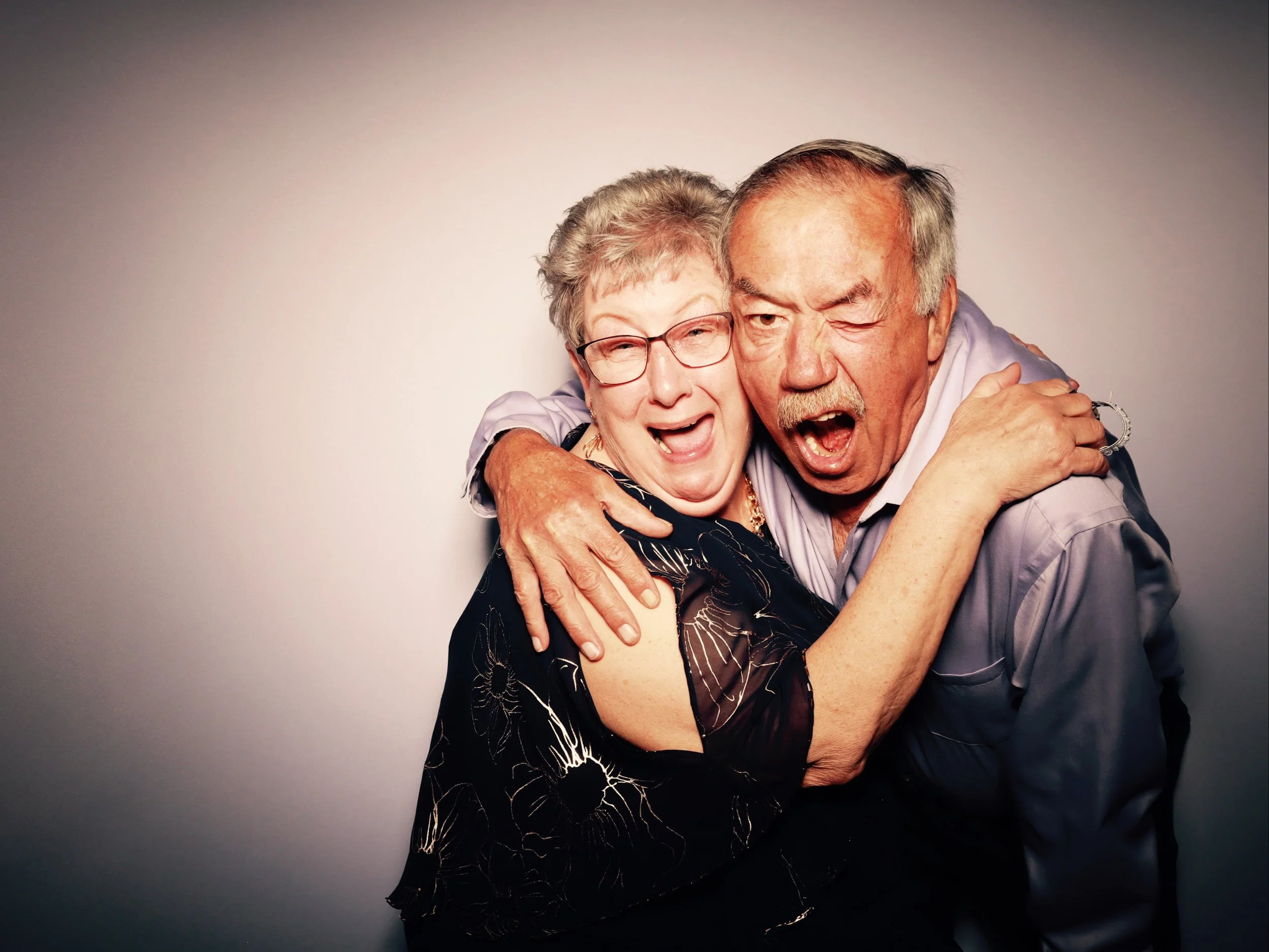 An elderly woman and man with gray hair and glasses are hugging and smiling, appearing joyful and affectionate.