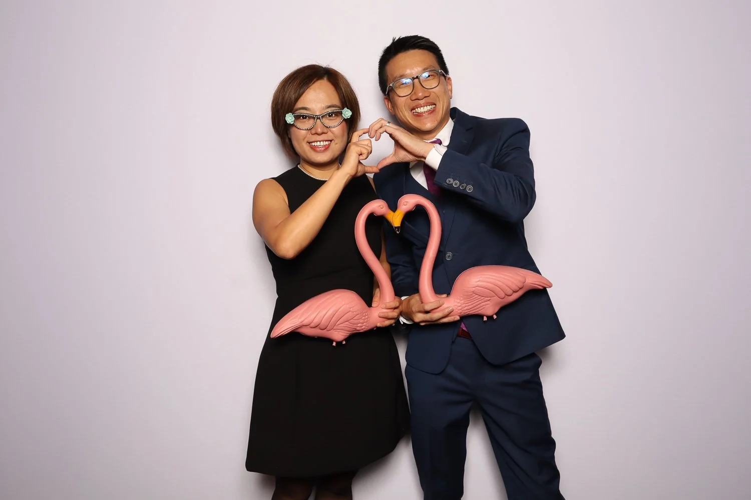 A smiling Asian couple in formal attire posing with pink plastic flamingo decorations, forming a heart shape with their hands against a plain white background.