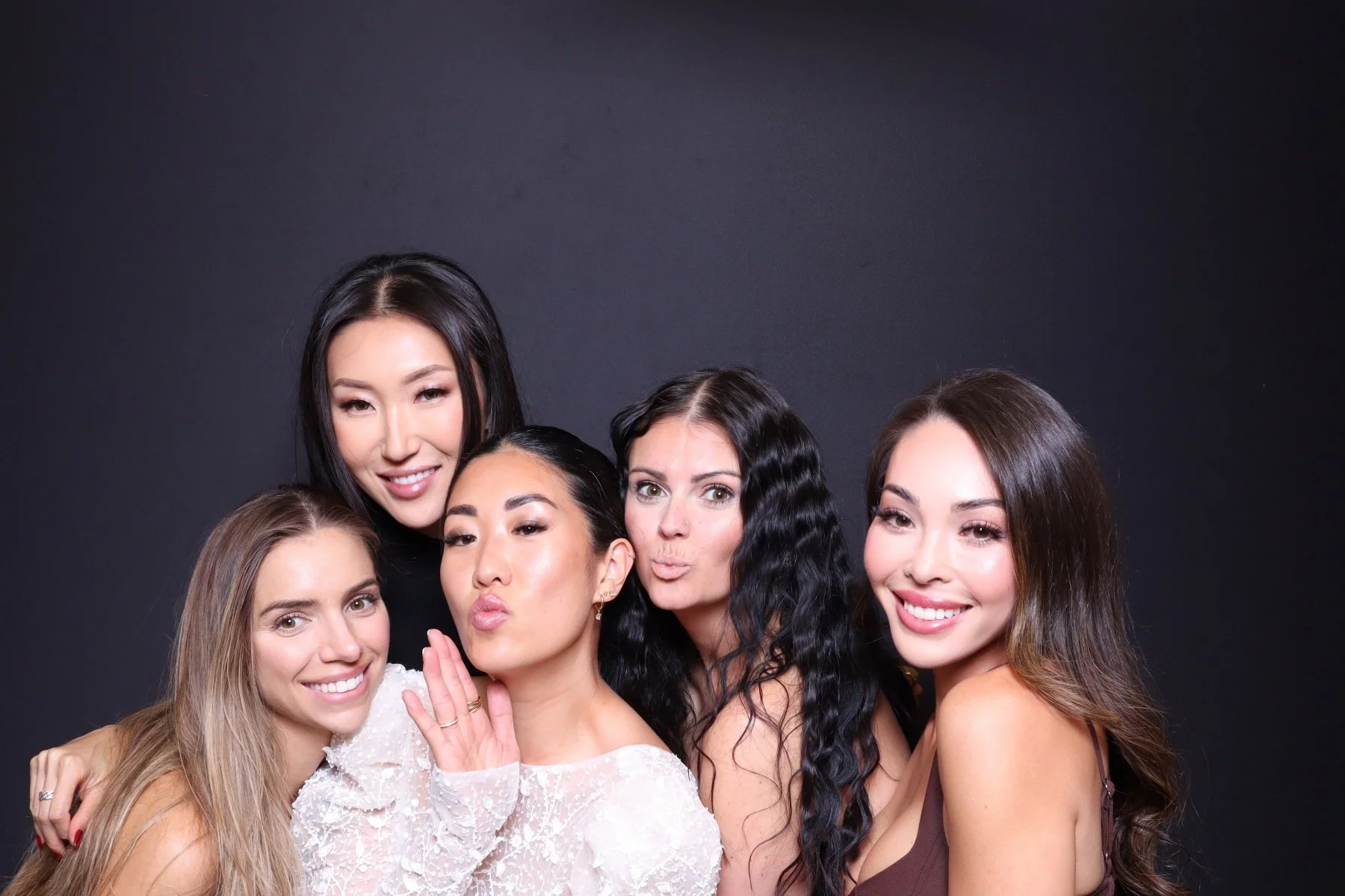 Five women posing together for a group photo against a black background, smiling and making playful expressions.