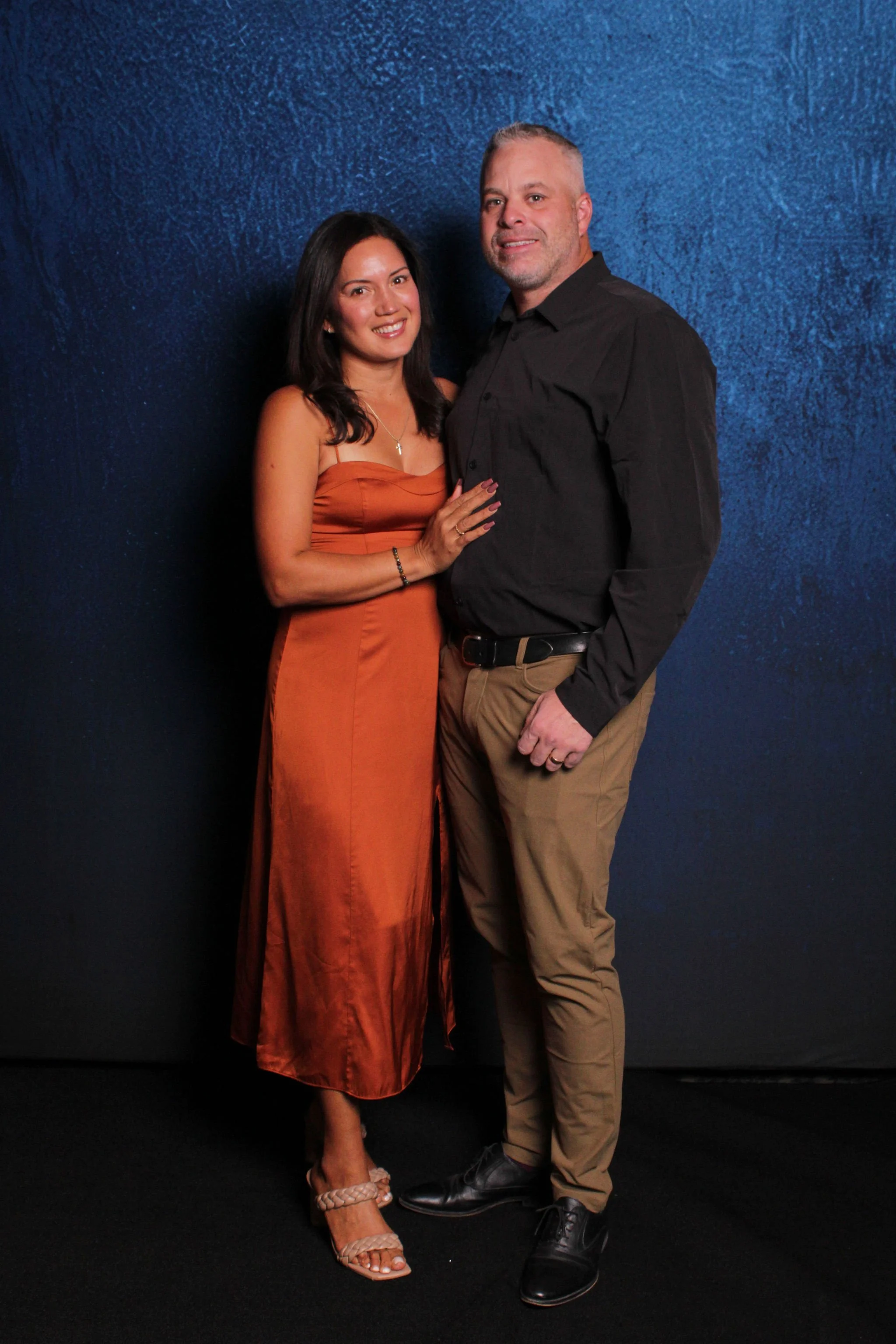 A man and woman standing close together, smiling, against a dark blue textured background.