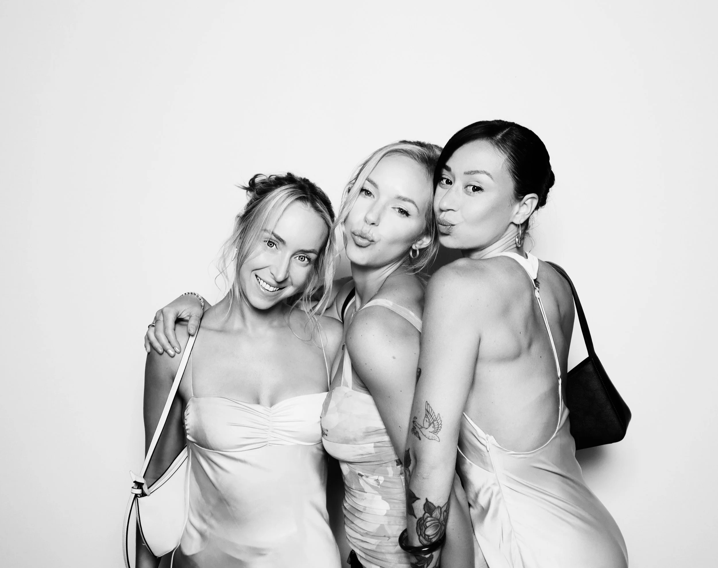 Three women dressed in light dresses, smiling and posing together for a photo against a plain background.