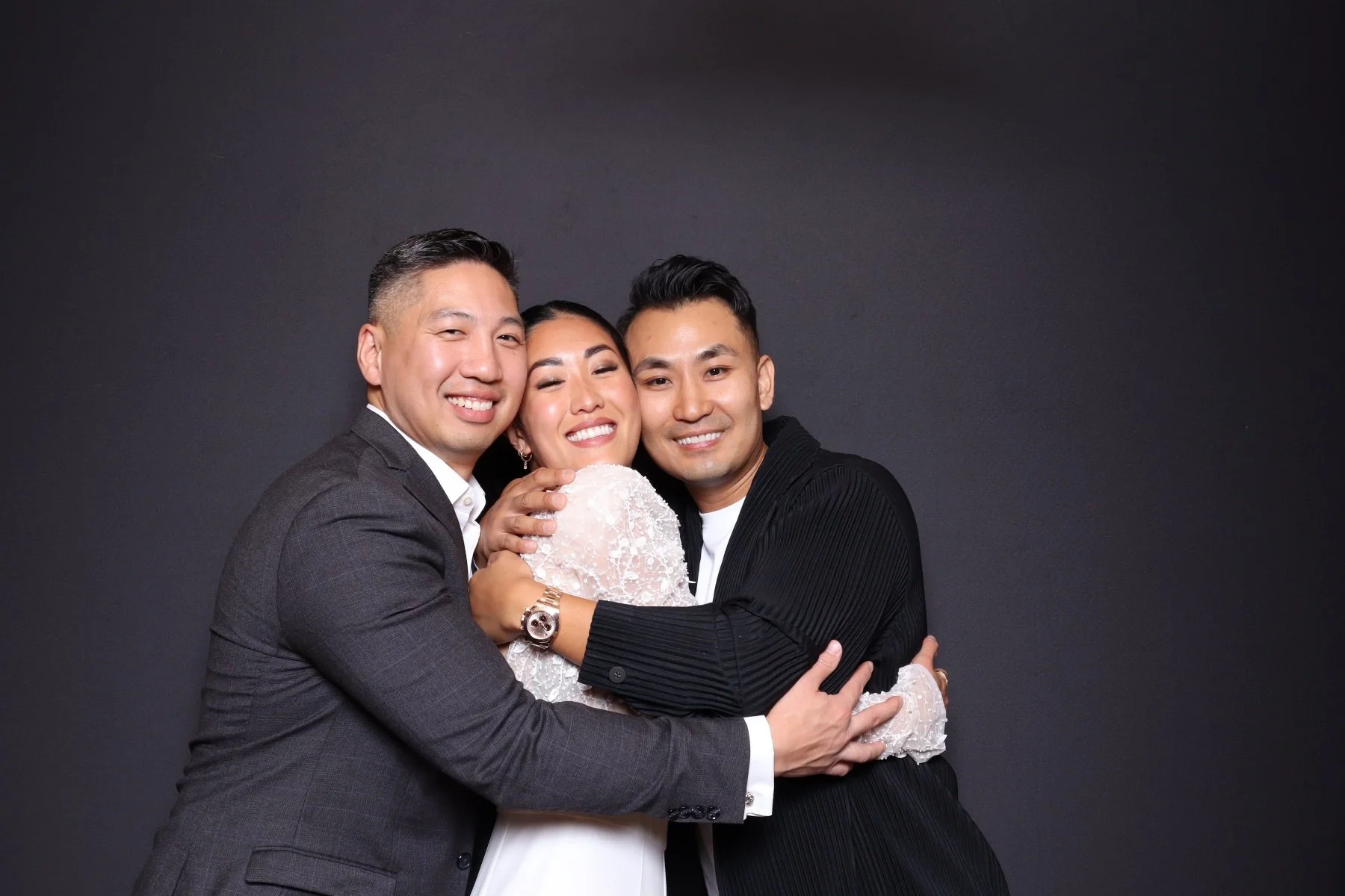 Three people smiling and hugging, standing against a dark background.