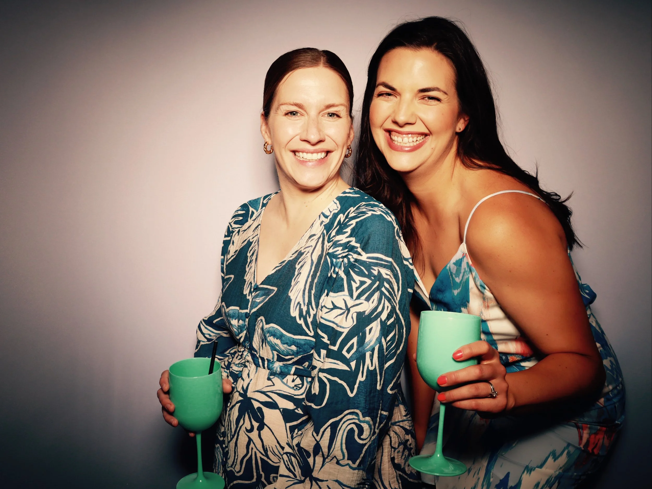 Two women smiling, holding green cocktails, standing close together against a neutral background.