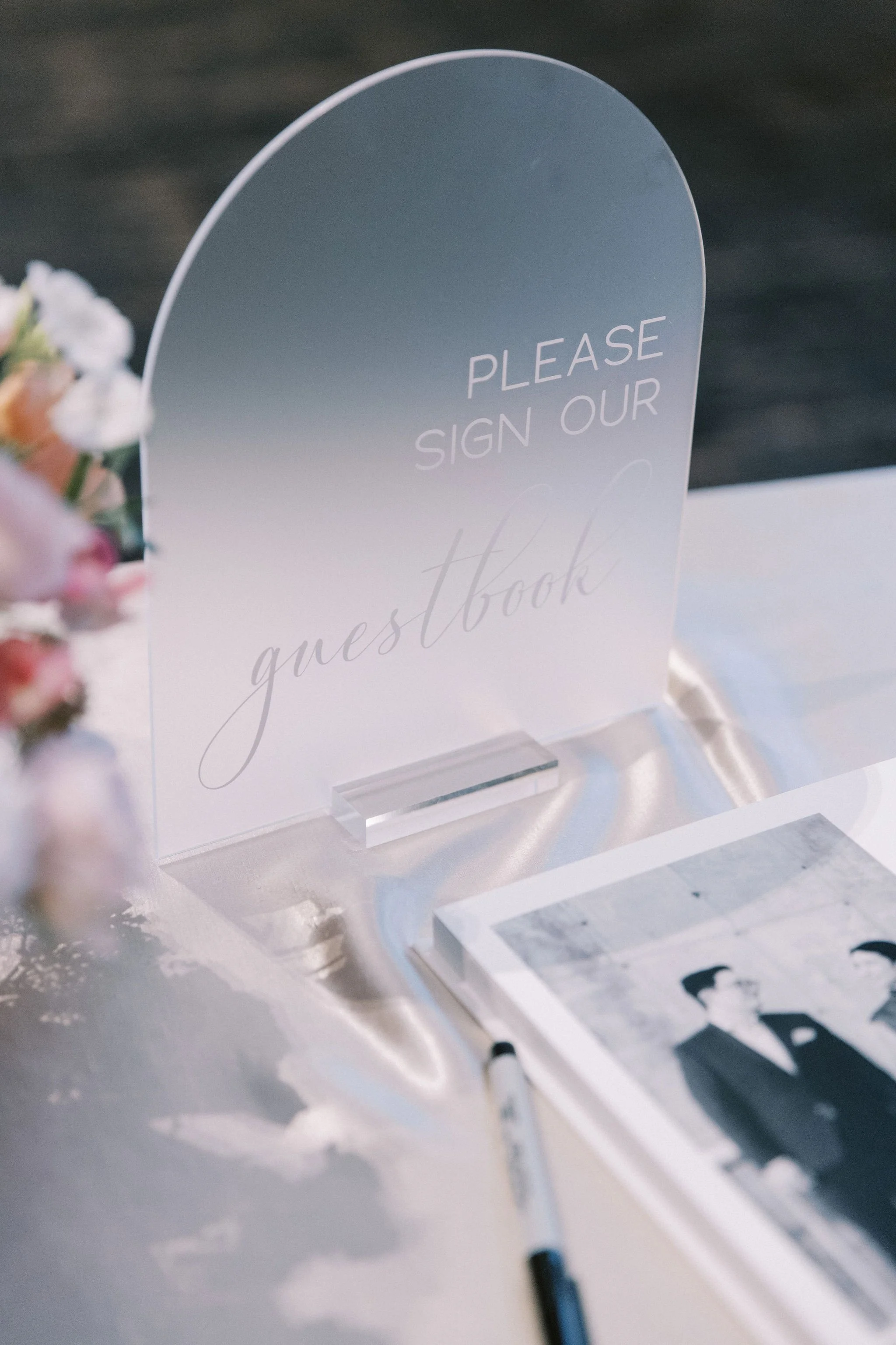 A transparent guestbook sign on a table at an event, with a pen nearby and a photograph of two people in formal attire.