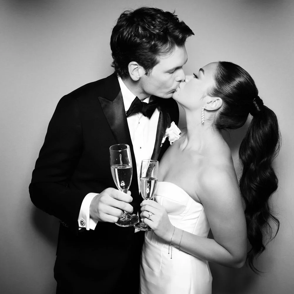 A black and white photo of a bride and groom sharing a kiss, each holding a champagne flute. The groom is wearing a tuxedo with a boutonniere, and the bride is in a strapless wedding dress with long earrings and a ponytail.