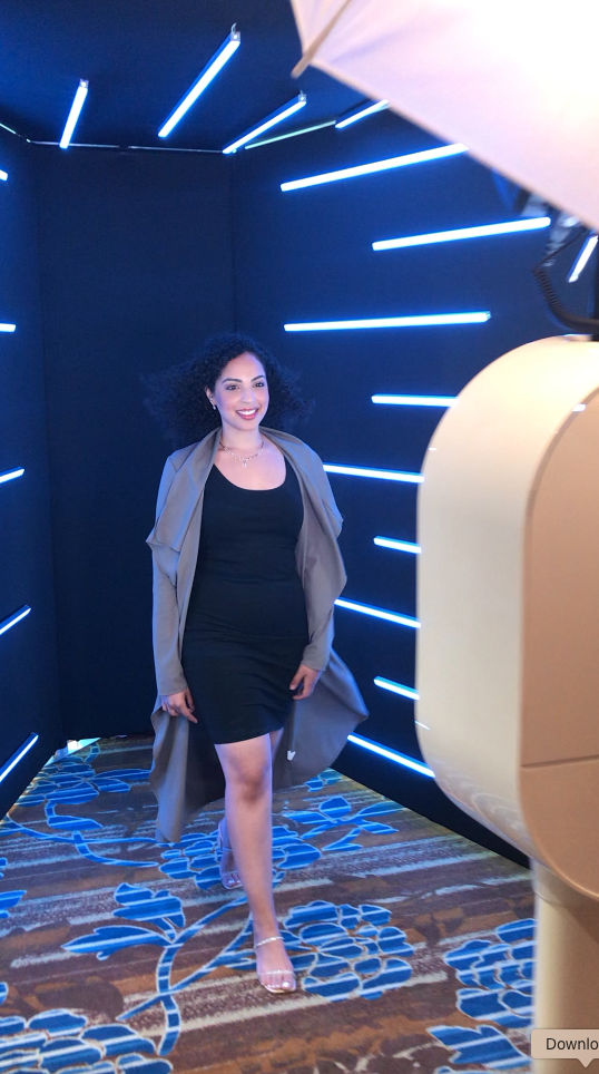 A woman in a black dress and gray coat standing in a room with blue neon strip lights on black walls, carpeted floor with blue floral pattern.