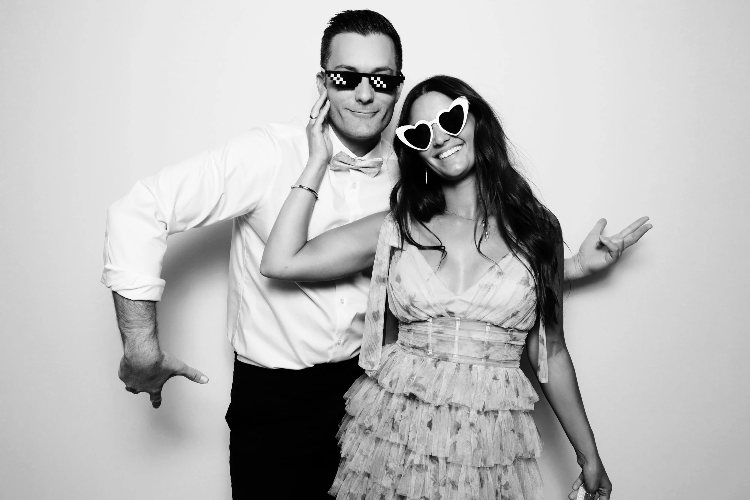 A man and woman dressed formally, wearing sunglasses, posing playfully in front of a plain wall.
