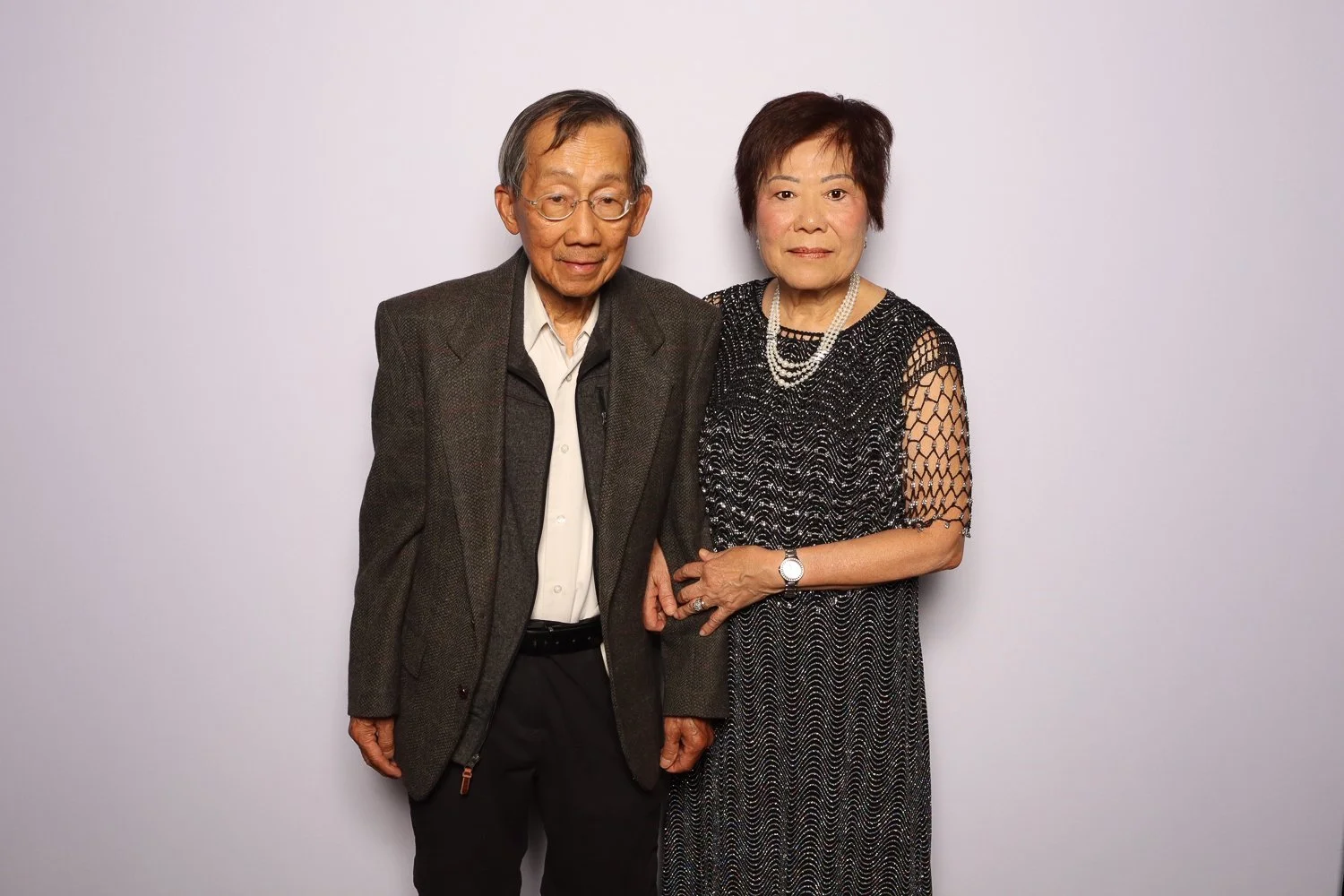 An elderly Asian couple standing together, dressed in formal attire, posing against a plain light-colored background.