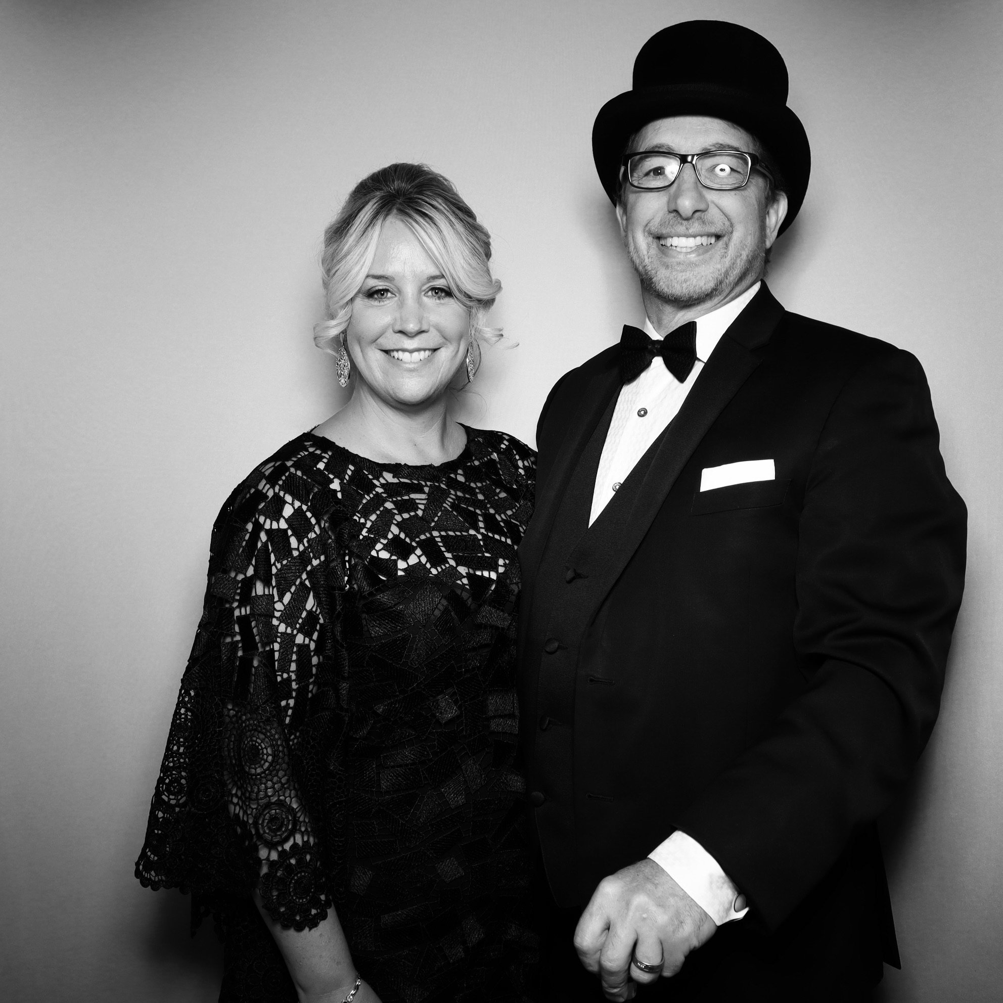 A man and woman dressed in formal attire posing for a photo, the man wearing a tuxedo with a bow tie and a top hat, and the woman wearing a lace dress, against a plain background.
