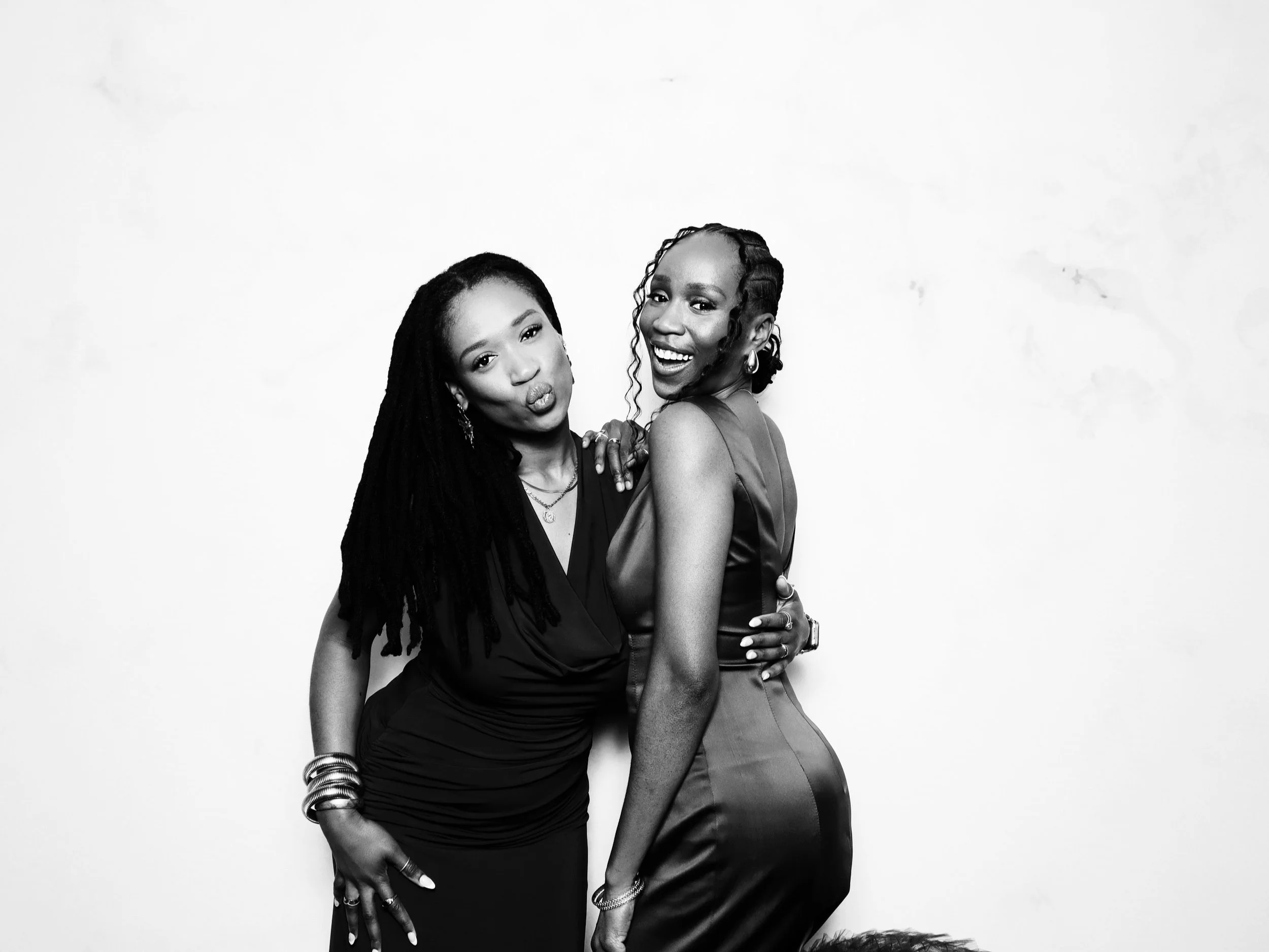 Two women in elegant dresses posing together against a white background, one making a pout and the other smiling.