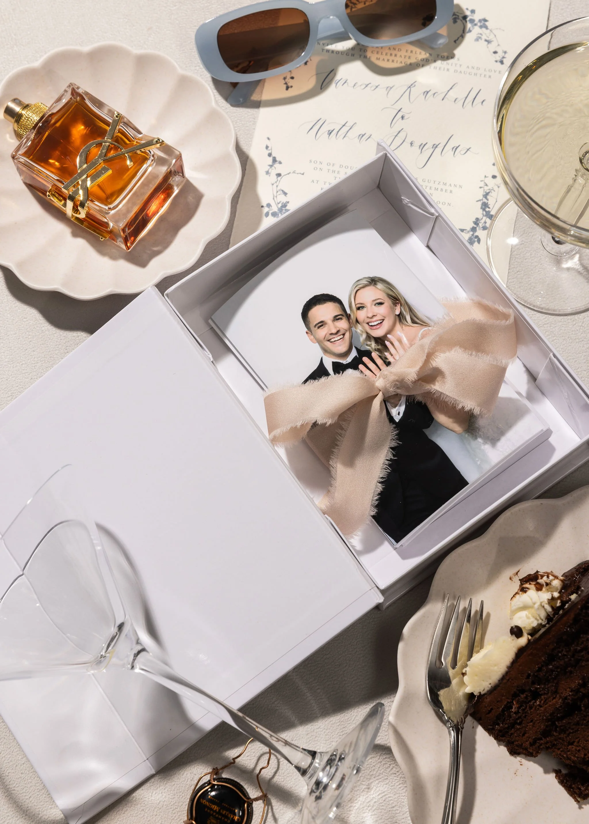 Wedding invitation, sunglasses, perfume bottle, champagne glass, wedding photo in box with beige ribbon, slice of chocolate cake with whipped cream, and empty wine glasses on a table.