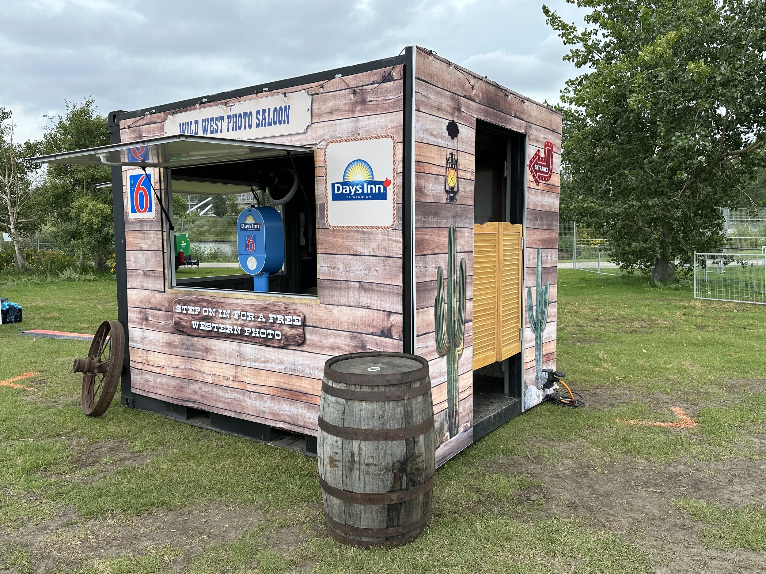 A small Western-themed photo booth with a rustic wood exterior, desert cacti decorations, and signs for a Western photo shoot. It has a window for taking photos, a barrel in front, and a sign inviting people to step in for a free Western photo.