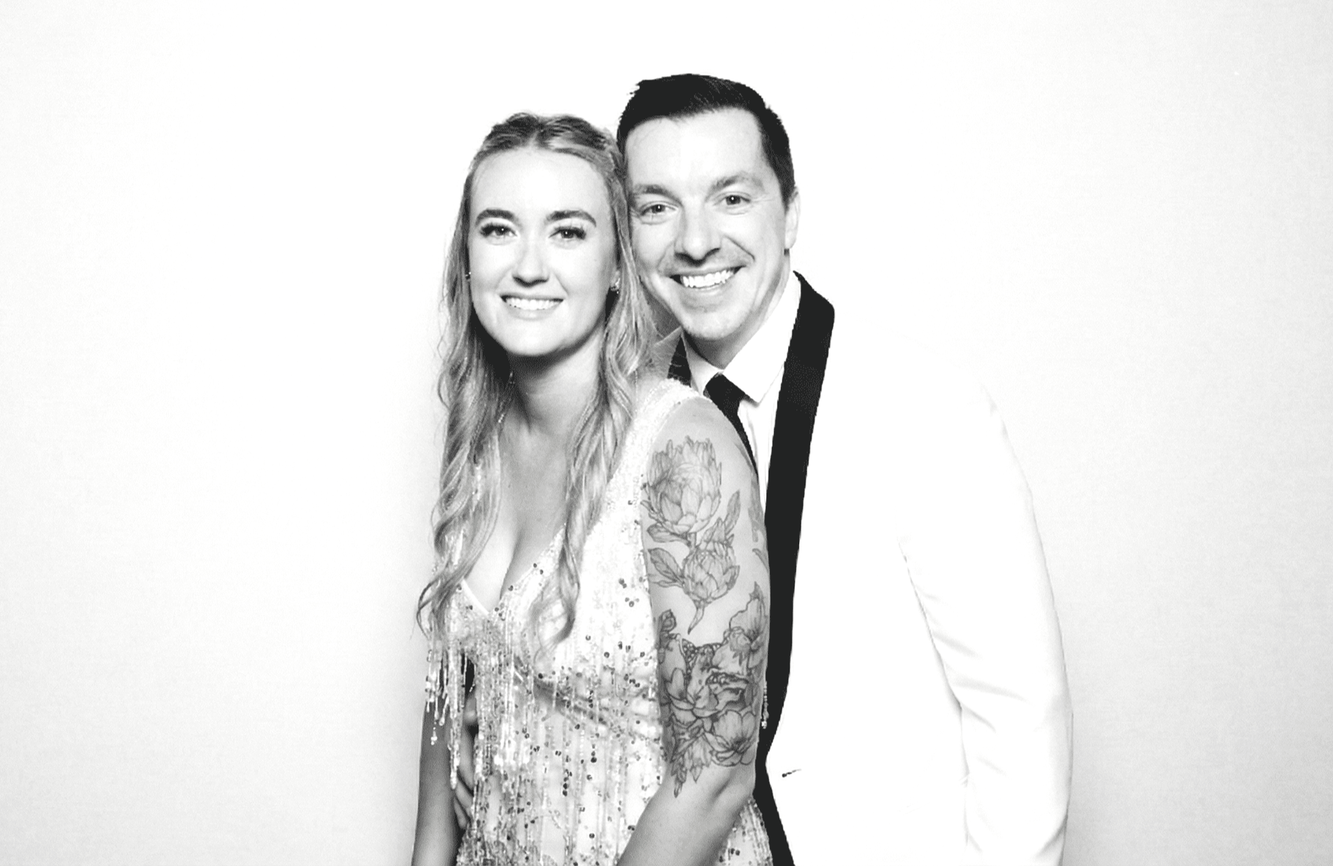 A black and white glam photo from a Flash Co. wedding photo booth featuring a smiling bride in a beaded gown and a groom in a white tuxedo jacket posing against a solid white backdrop.