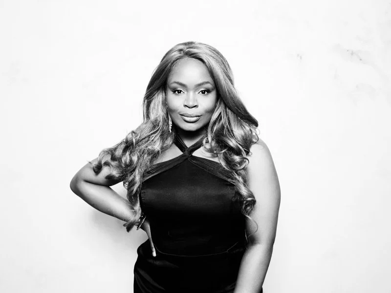 Black and white portrait of a woman with long wavy hair, wearing a sleeveless dress, standing against a plain background with one hand on her hip.