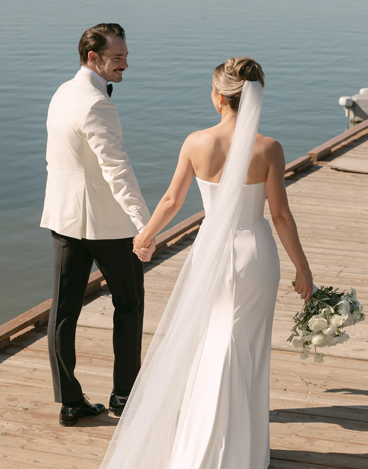 A bride and groom holding hands on a wooden dock by the water, with the bride holding a bouquet of white flowers.