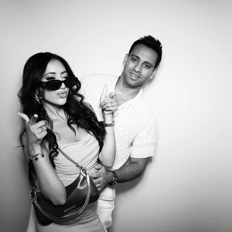 A woman with long dark hair wearing sunglasses, a light dress, and jewelry, standing next to a man with short dark hair in a casual shirt. They are posing playfully against a plain background.