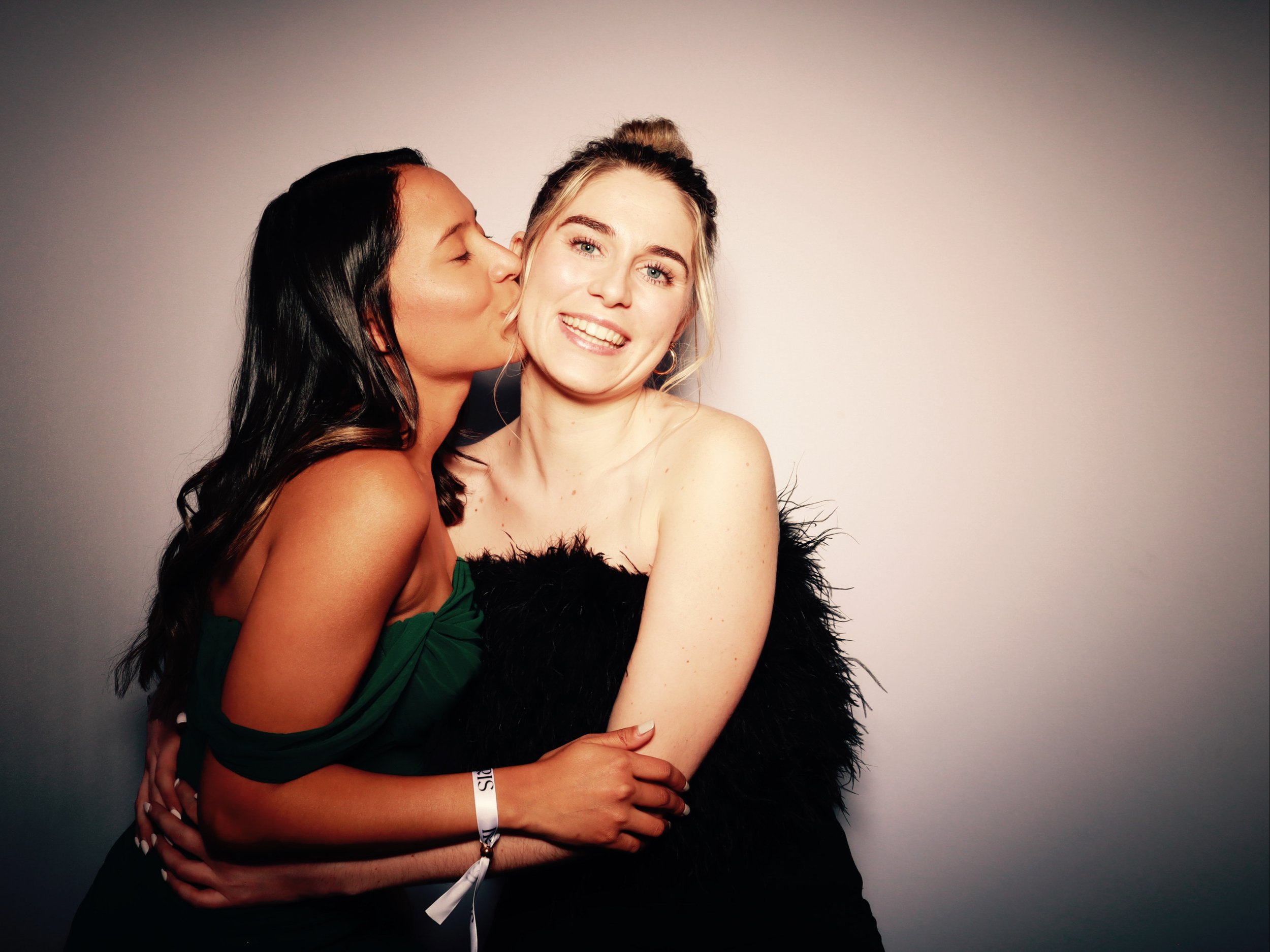 Two women hugging and smiling, one kissing the other's cheek in a friendly gesture, against a plain background.