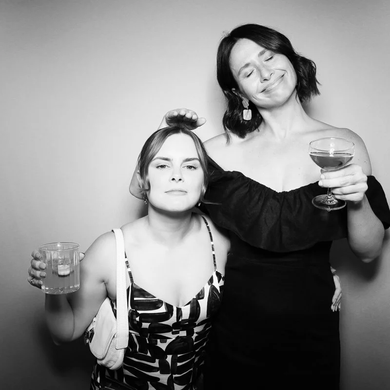 Two women at a party holding drinks, with one making a playful face and the other smiling with eyes closed.