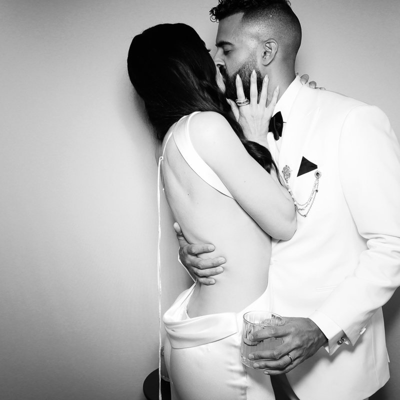 A black-and-white photo of a couple kissing, with the woman wearing a sleeveless dress and the man in a white suit with a black bow tie. The woman's hand with a ring touches the man's face, and he holds a glass of drink in his hand.
